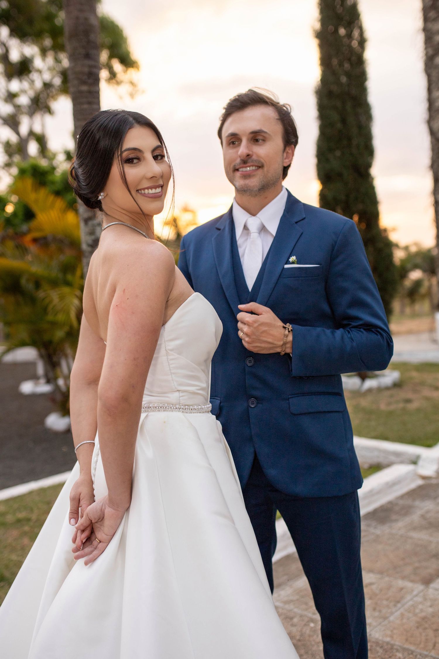 Bride in white strapless gown and groom in navy blue suit hold hands at outdoor sunset wedding ceremony.