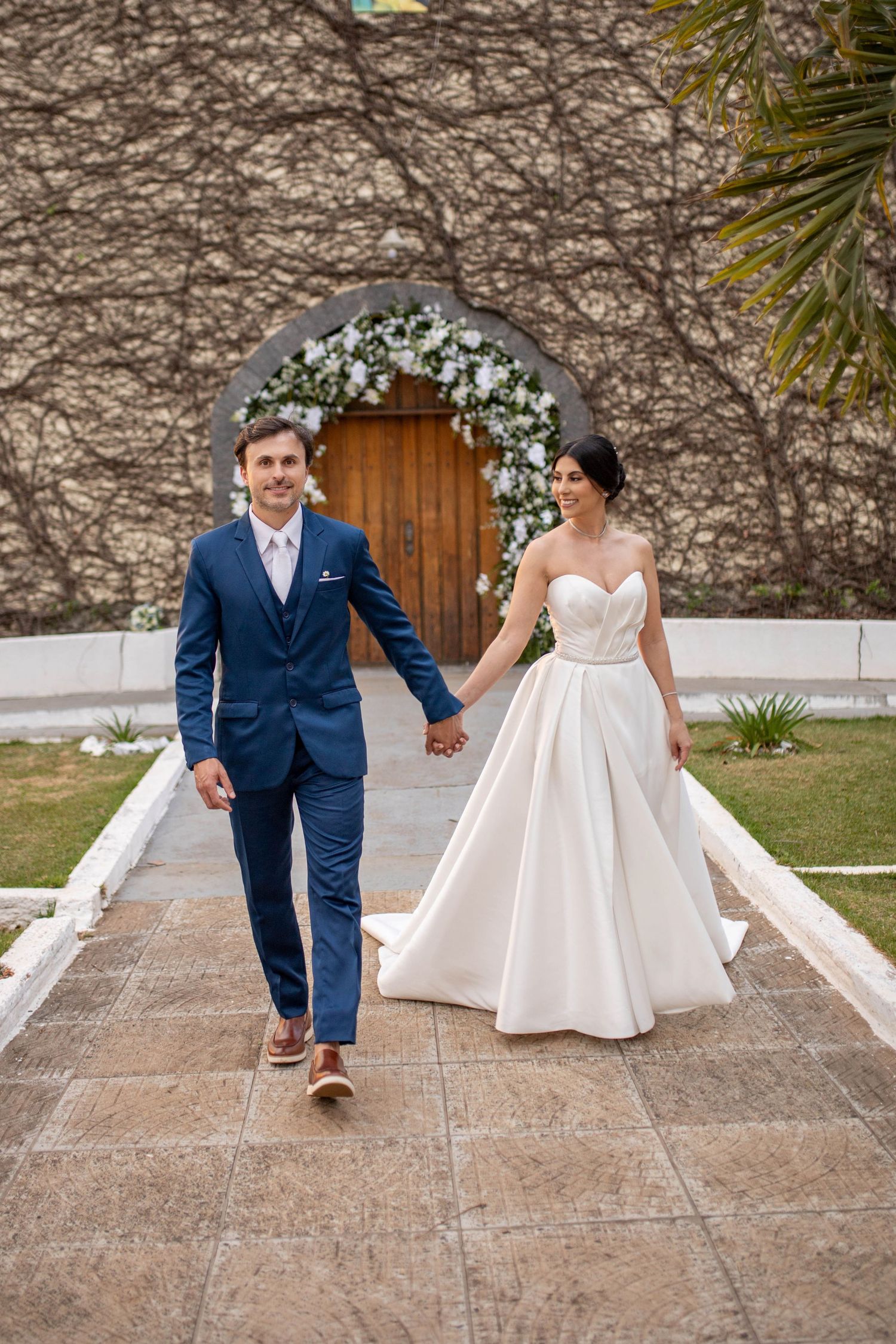 Bride in white strapless ballgown and groom in blue suit hold hands walking from floral-adorned wooden church doors.