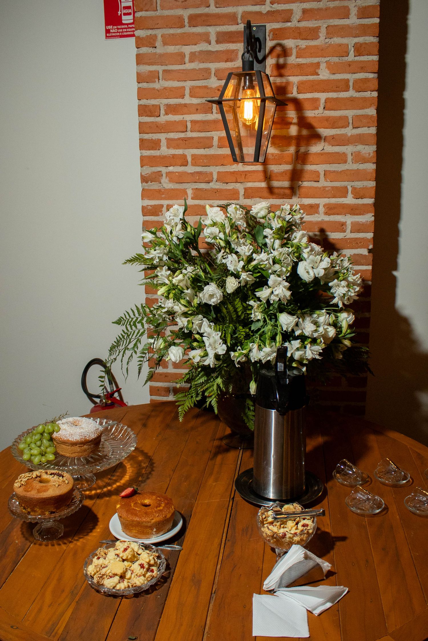 Elegant wooden table with white flower bouquet, pastries, and grapes beside a brick wall with vintage lantern.