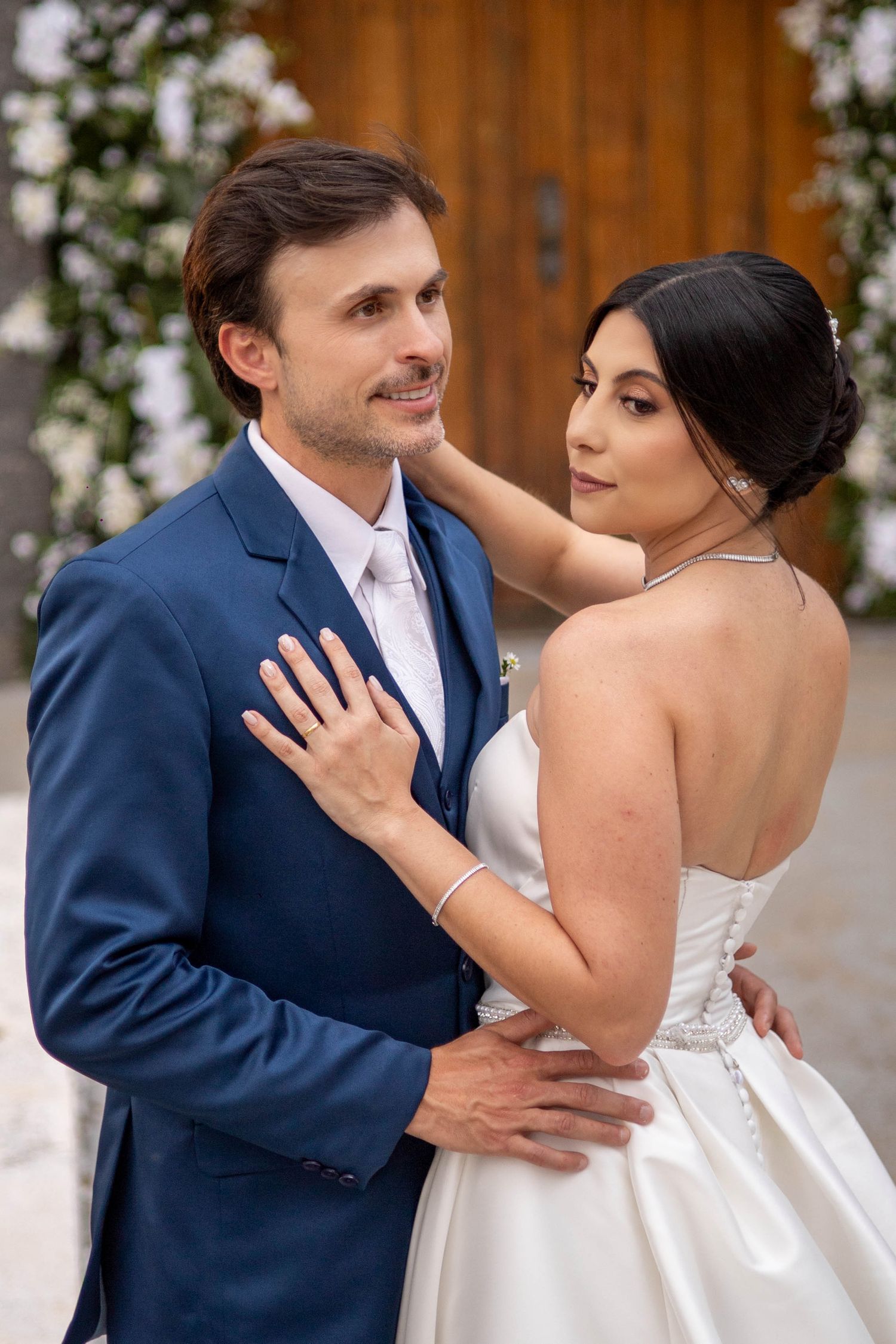 Bride in white strapless gown and groom in blue suit share romantic embrace at elegant floral wedding ceremony.