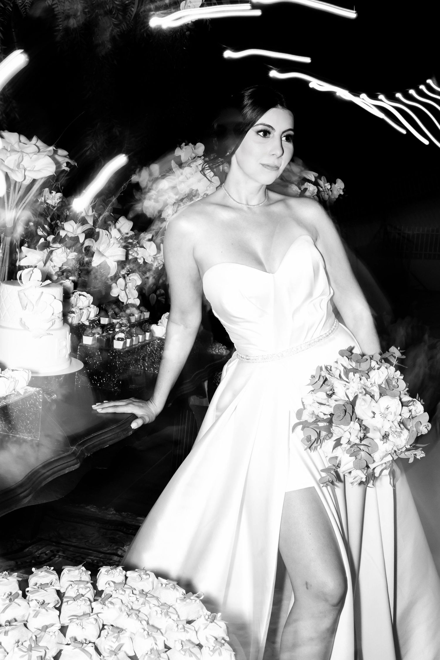 Black and white photo of a bride in a strapless gown holding a bouquet near a wedding cake.