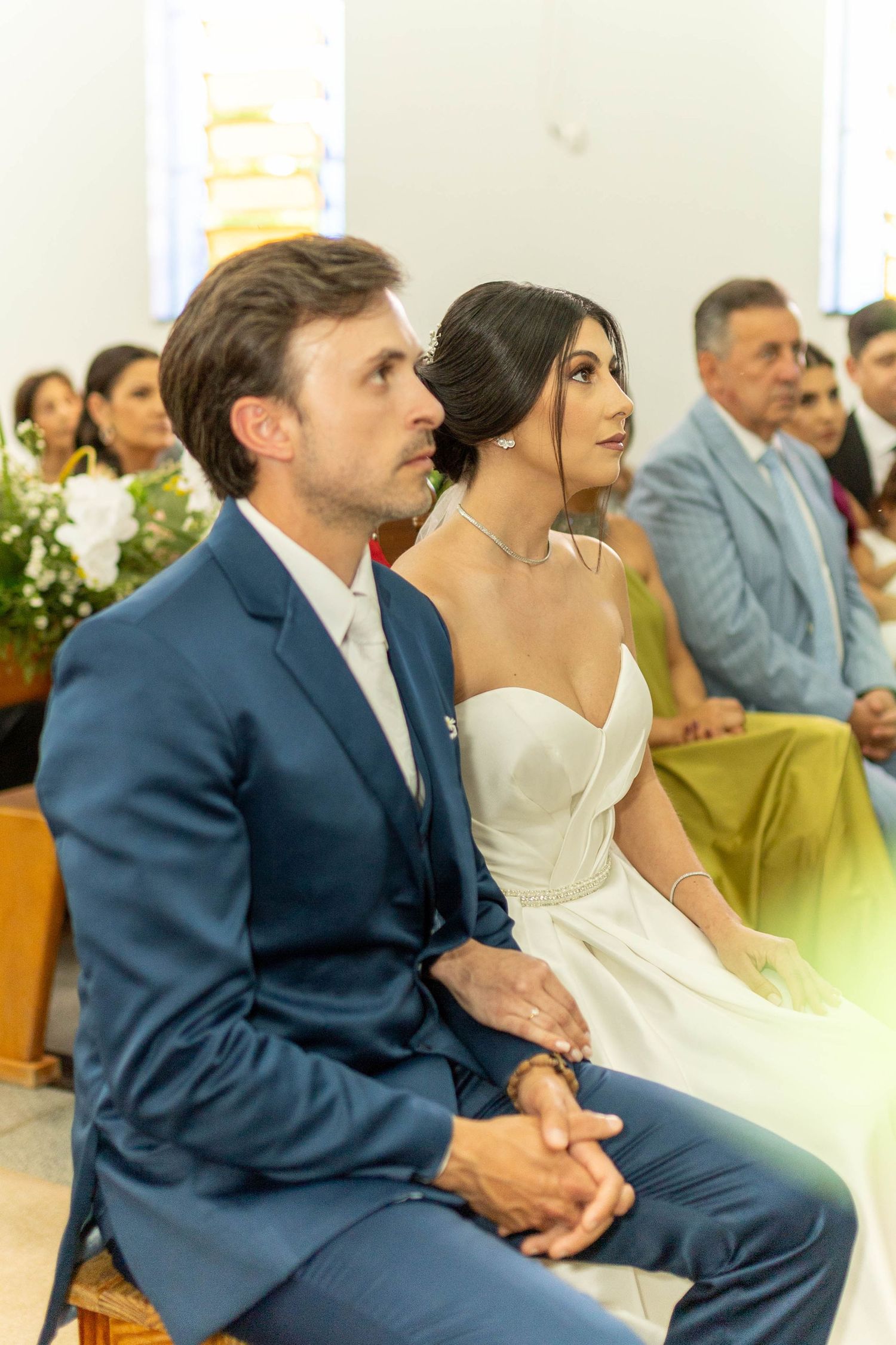 Bride and groom sitting together during wedding ceremony in church, groom in blue suit, bride in white strapless gown.
