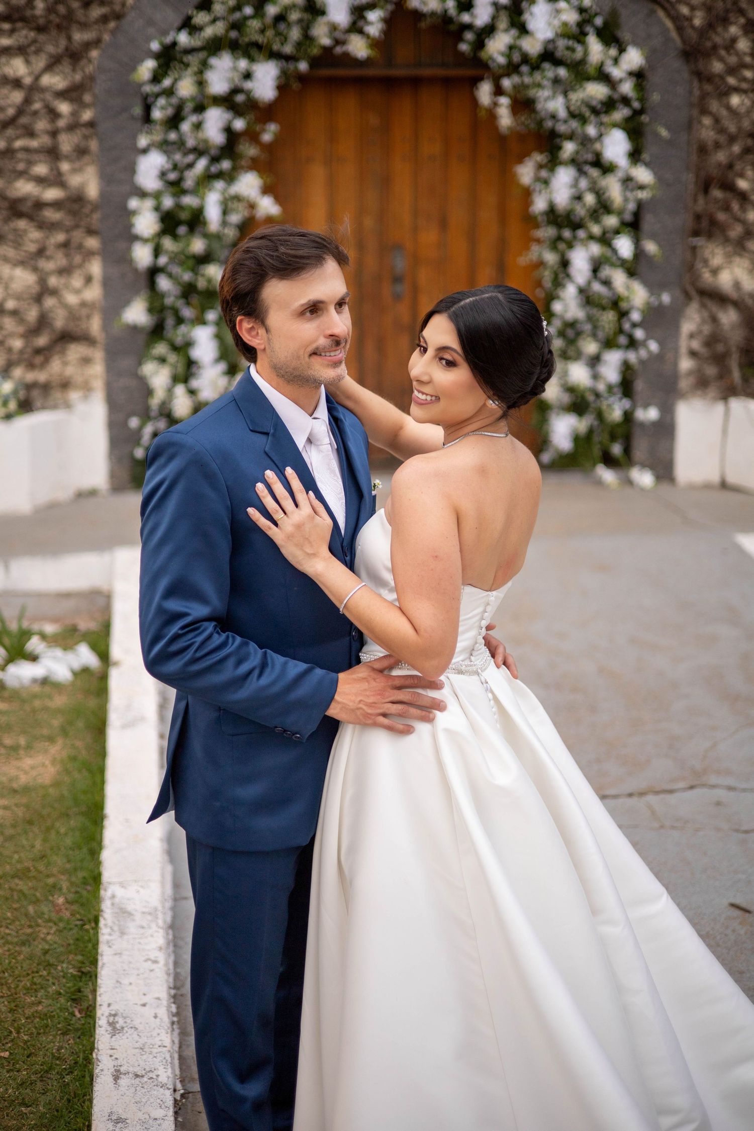 Bride and groom embrace before floral arch at church wedding, bride in strapless gown, groom in navy suit.