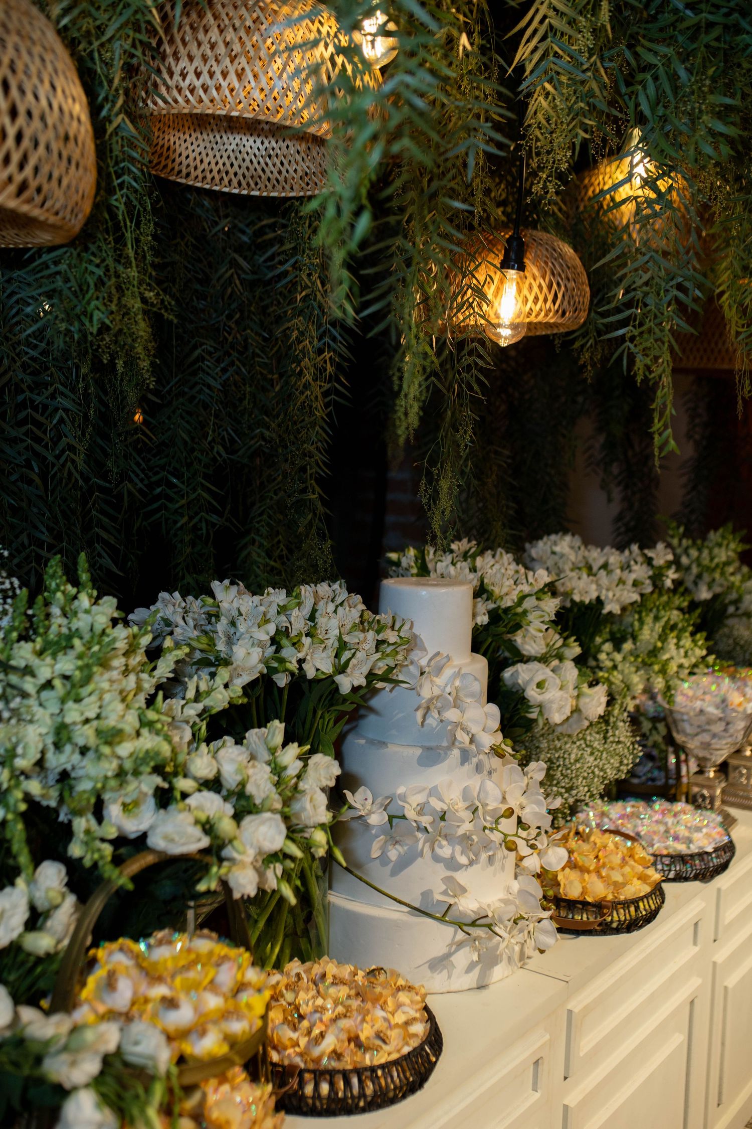Elegant wedding dessert table with white tiered cake, fresh white flowers, assorted sweets, and warm hanging lights amid lush greenery.