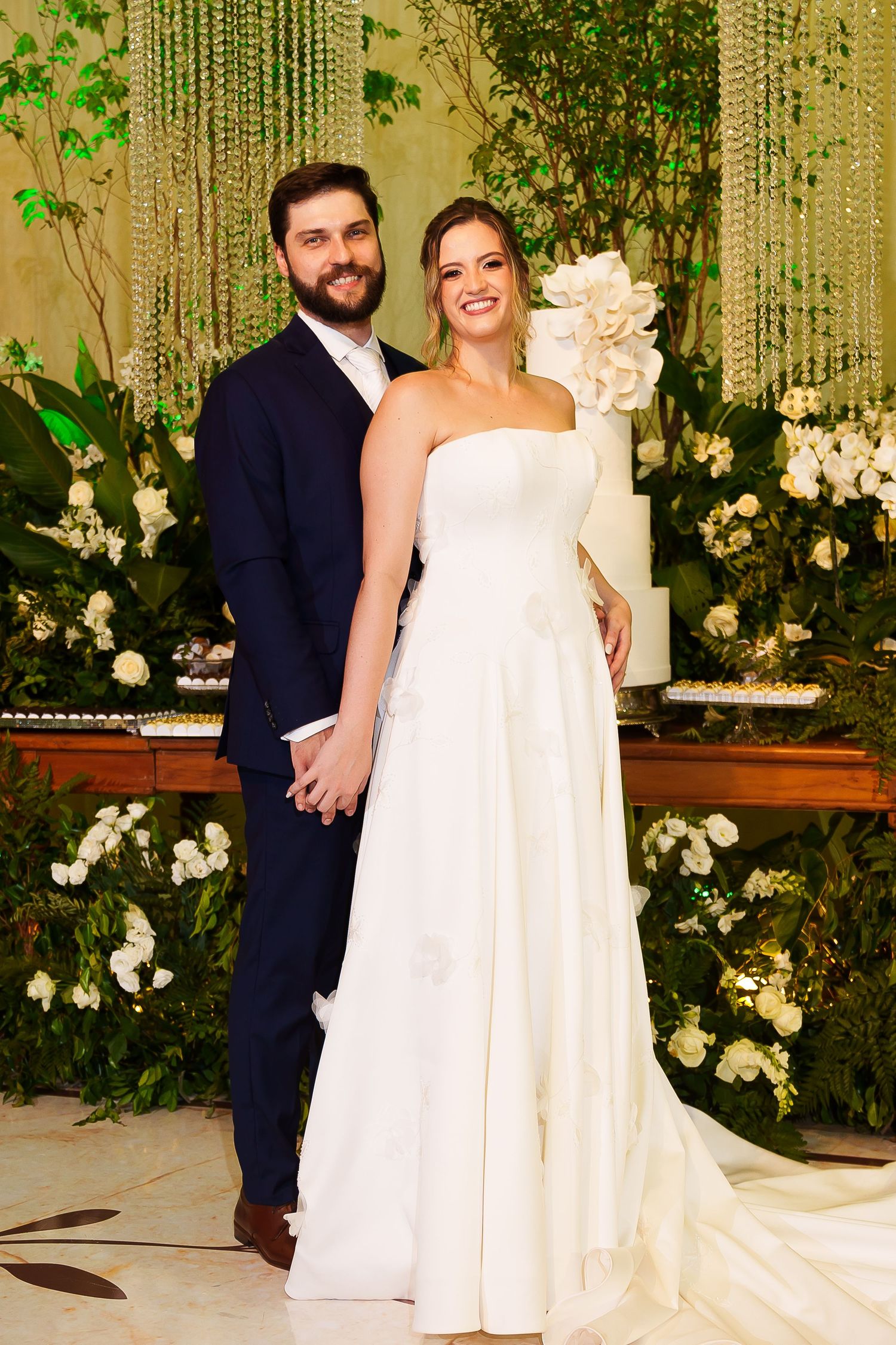 Bride in white strapless gown with floral detail stands with groom in navy suit at elegant wedding reception.