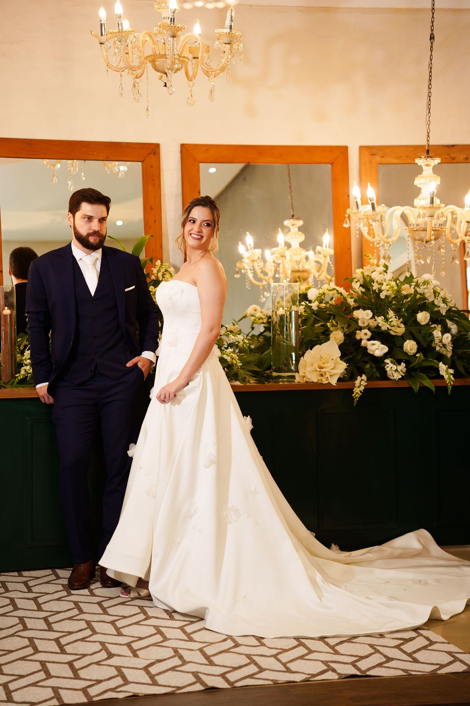 Bride in white strapless gown and groom in navy suit pose elegantly in a candlelit wedding venue with floral arrangements.