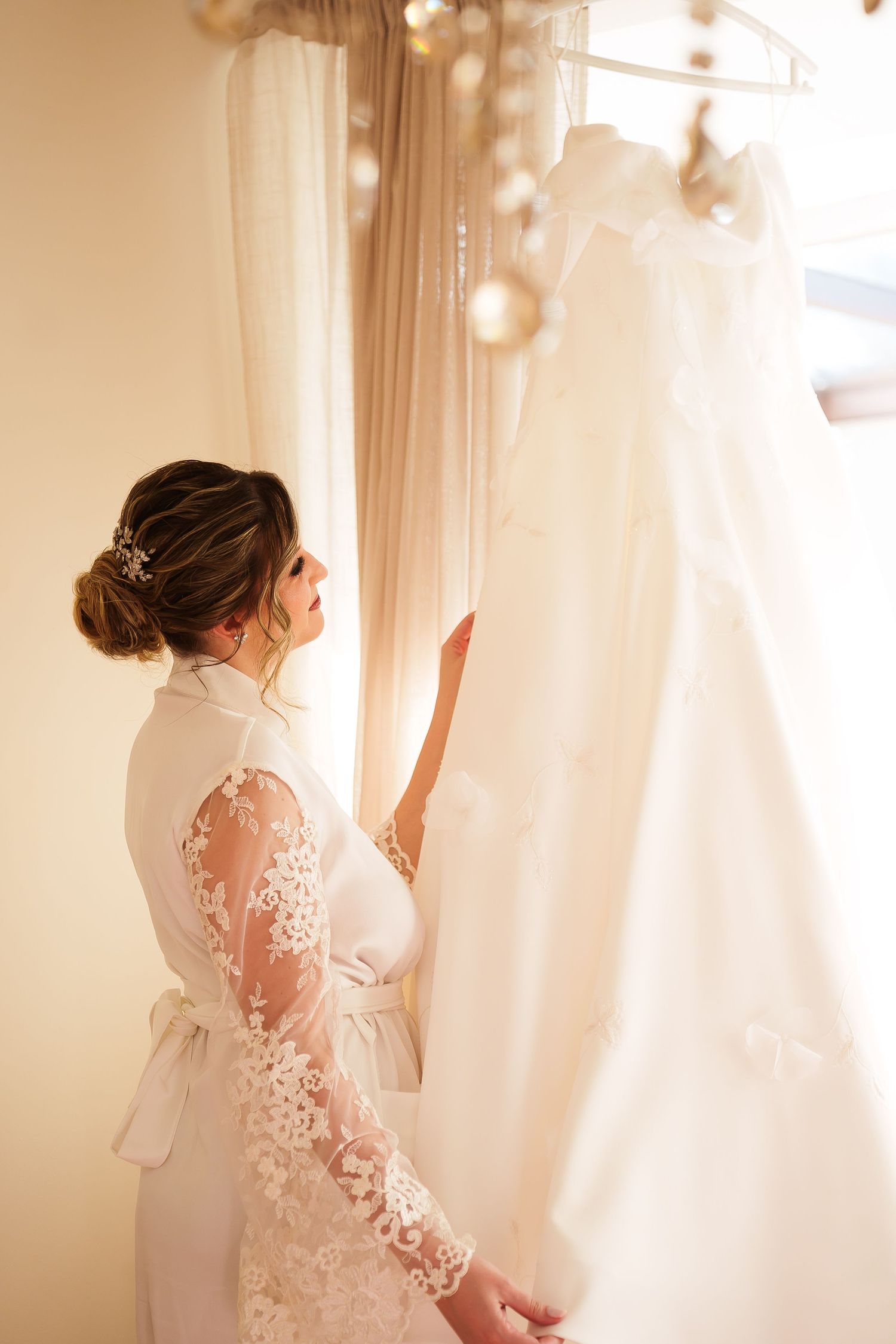 Bride in lace robe admiring her wedding dress on a hanger near a window with soft warm lighting.