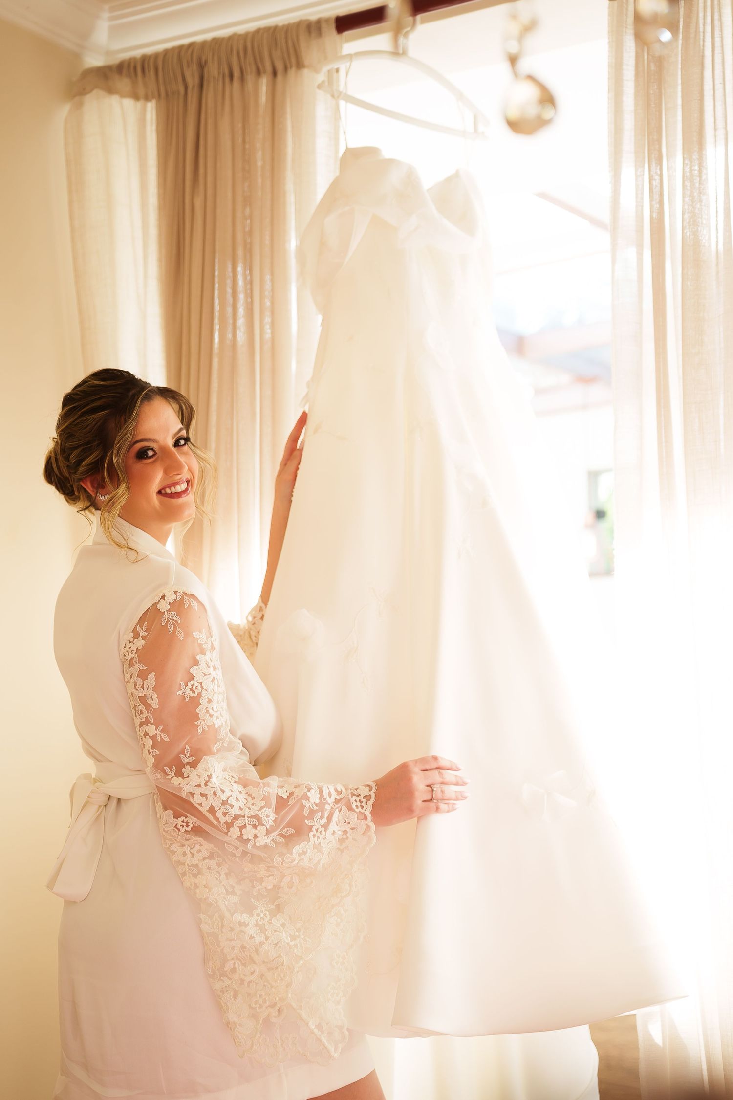 Smiling bride in lace robe holding her white wedding dress near a sunlit window before the ceremony.