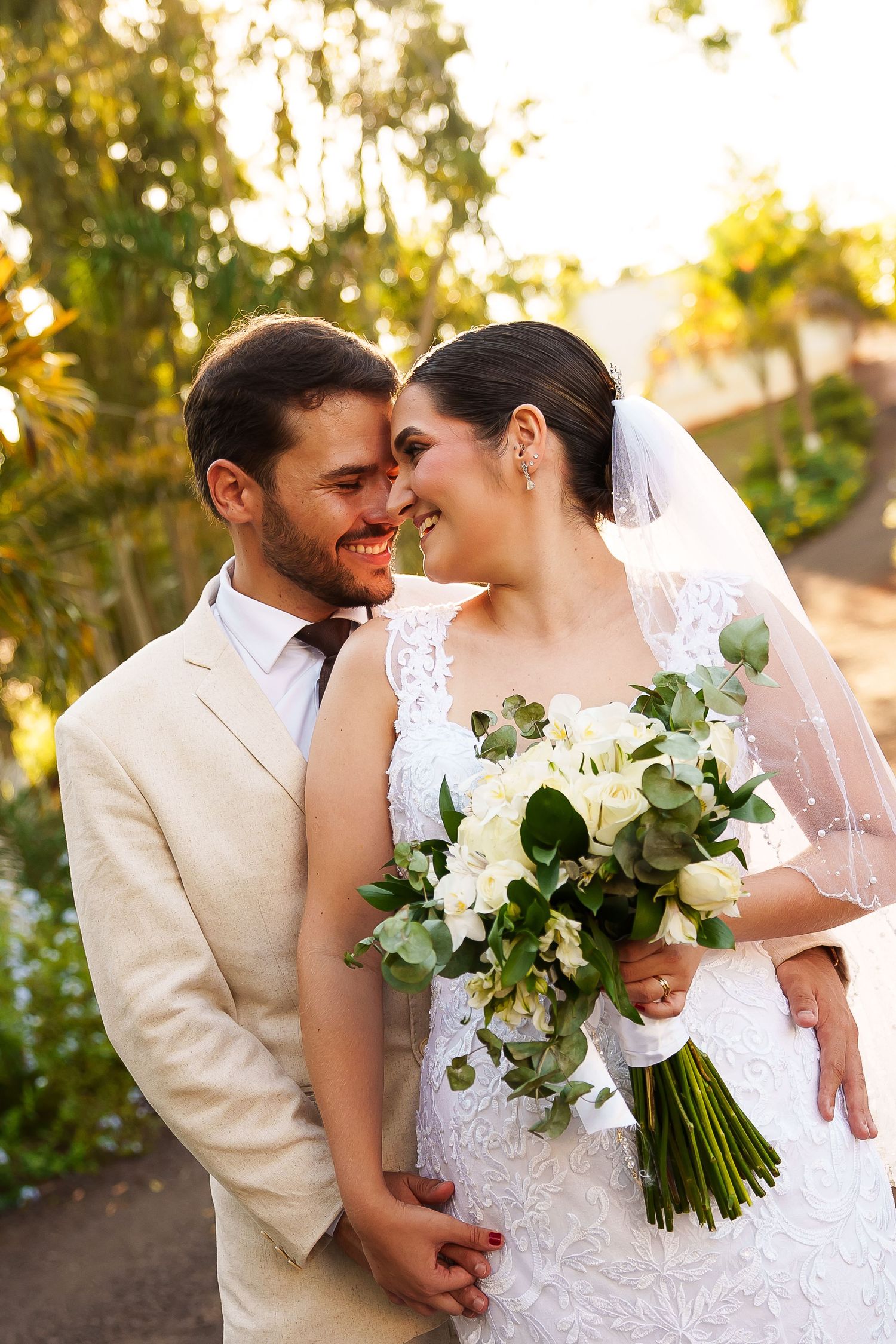Smiling bride and groom touch foreheads outdoors, bride holding white floral bouquet with eucalyptus in golden sunlight.