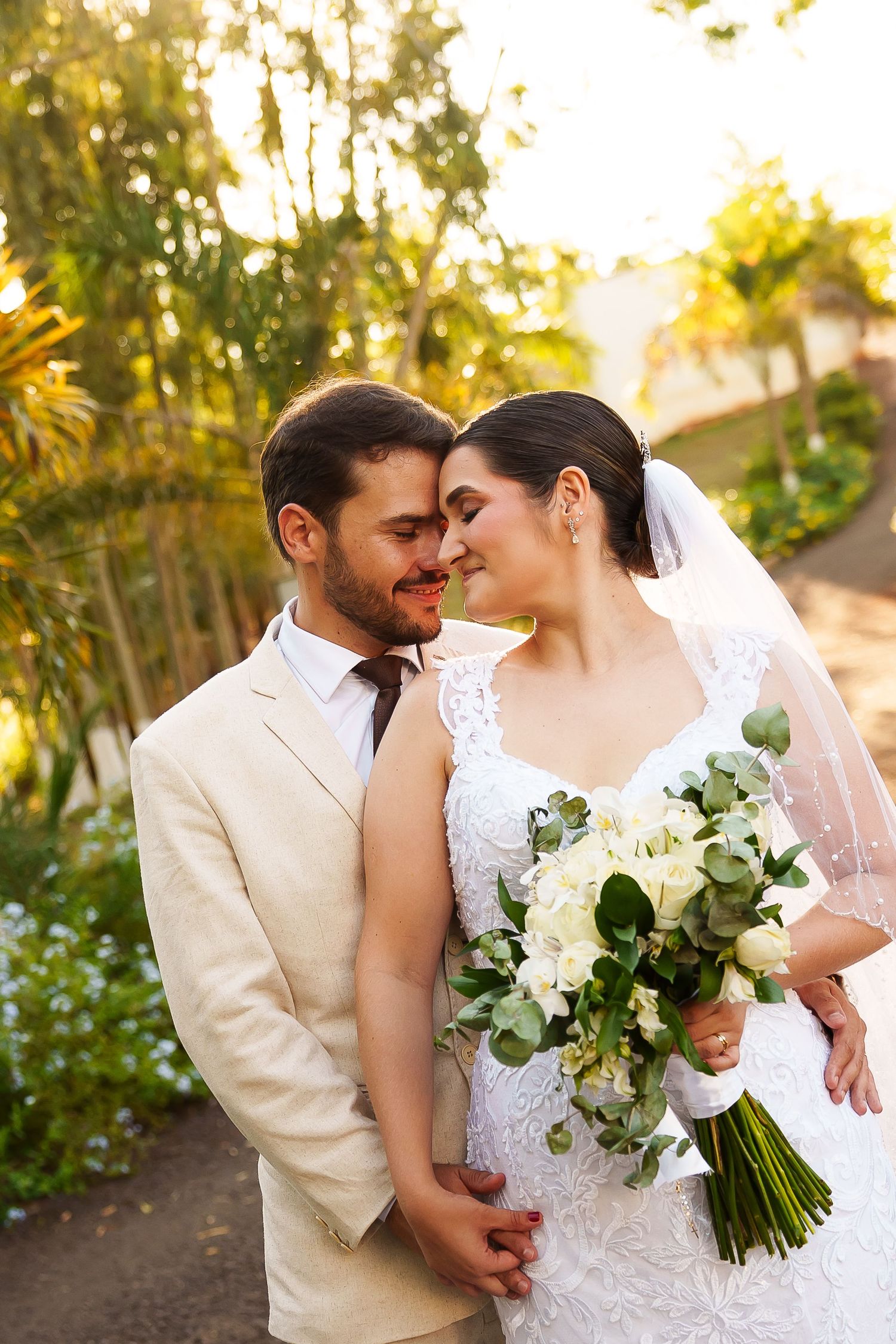 A bride and groom share a romantic forehead touch outdoors, she holds white roses bouquet, he wears beige suit.