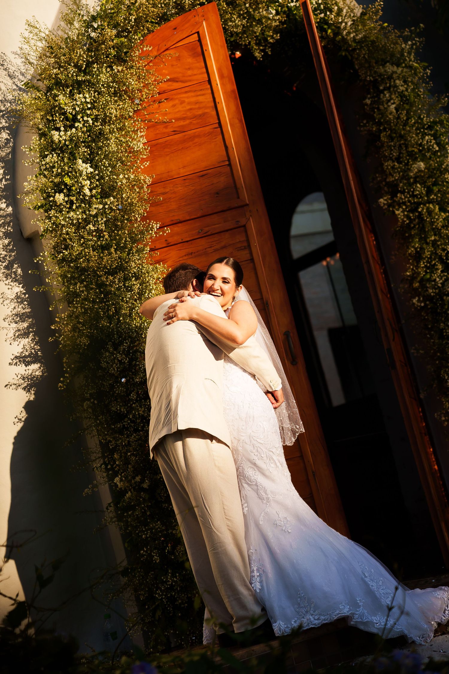 Bride and groom embracing in front of ornate wooden church doors surrounded by lush greenery on their wedding day.