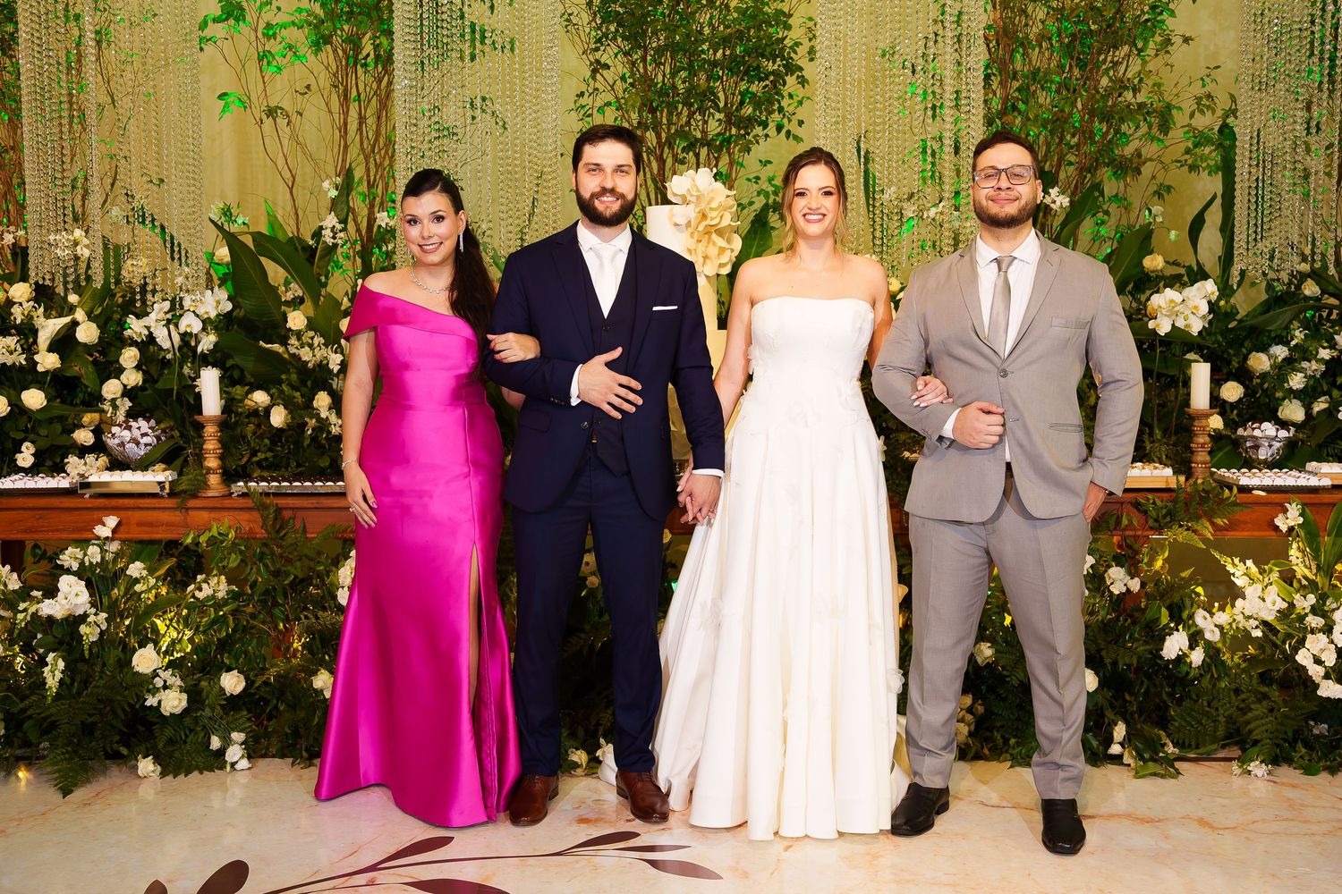 Four elegantly dressed guests pose together at a wedding reception with floral decorations in the background.