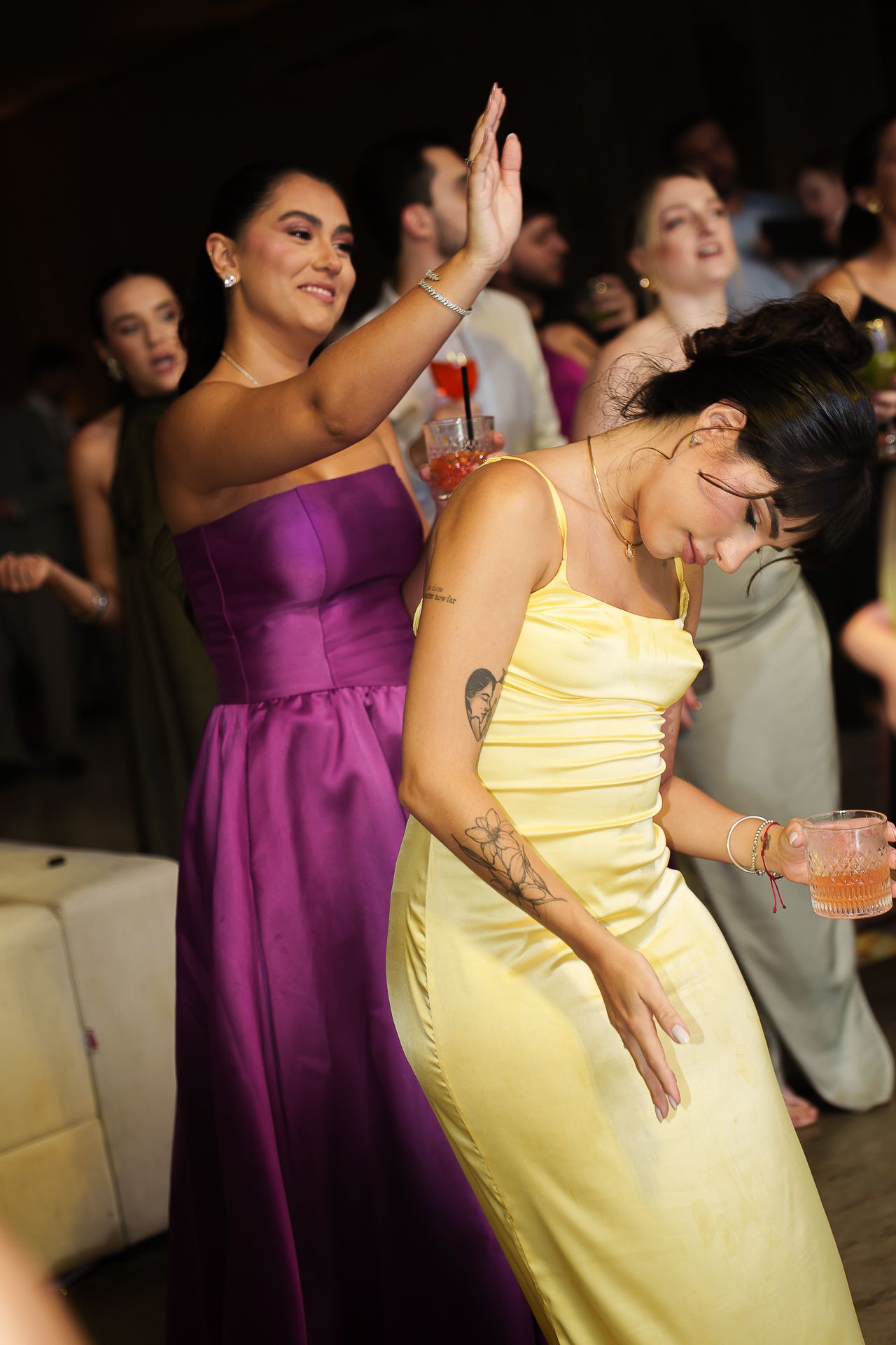 Two women in elegant satin gowns dancing joyfully at a formal event, one in purple and one in yellow.