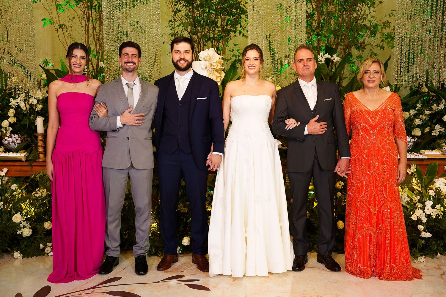 Wedding party of six posing elegantly, bride in white gown, guests in formal attire, lush green floral backdrop.