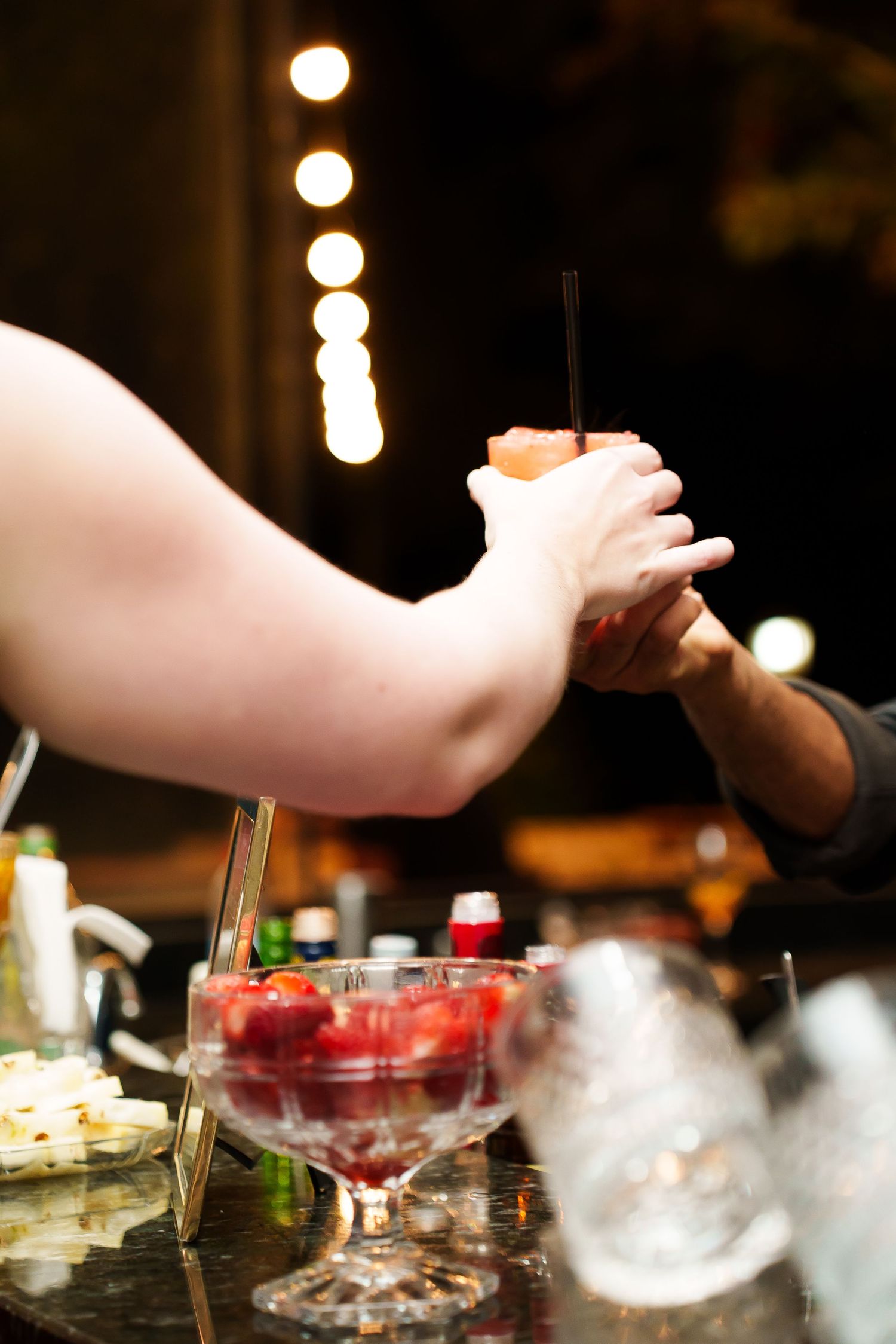 Two bartenders collaborate over a cocktail bar with fresh strawberries in a glass bowl and bokeh lights in background.
