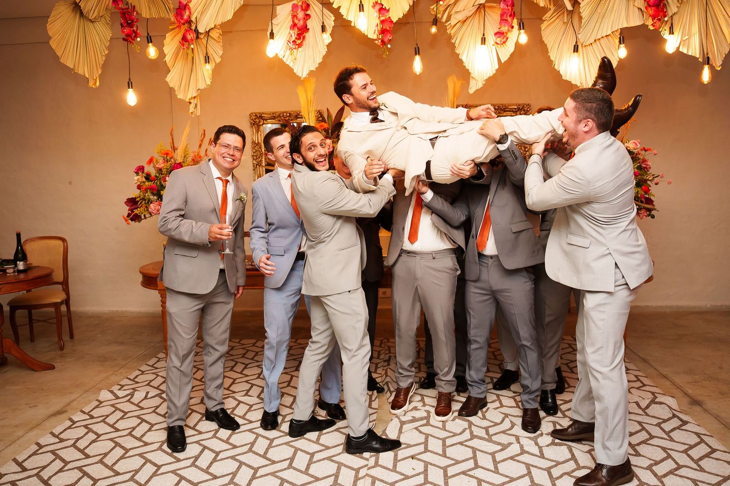 Groomsmen in gray suits with orange ties lift the groom in celebration at a festive wedding reception.