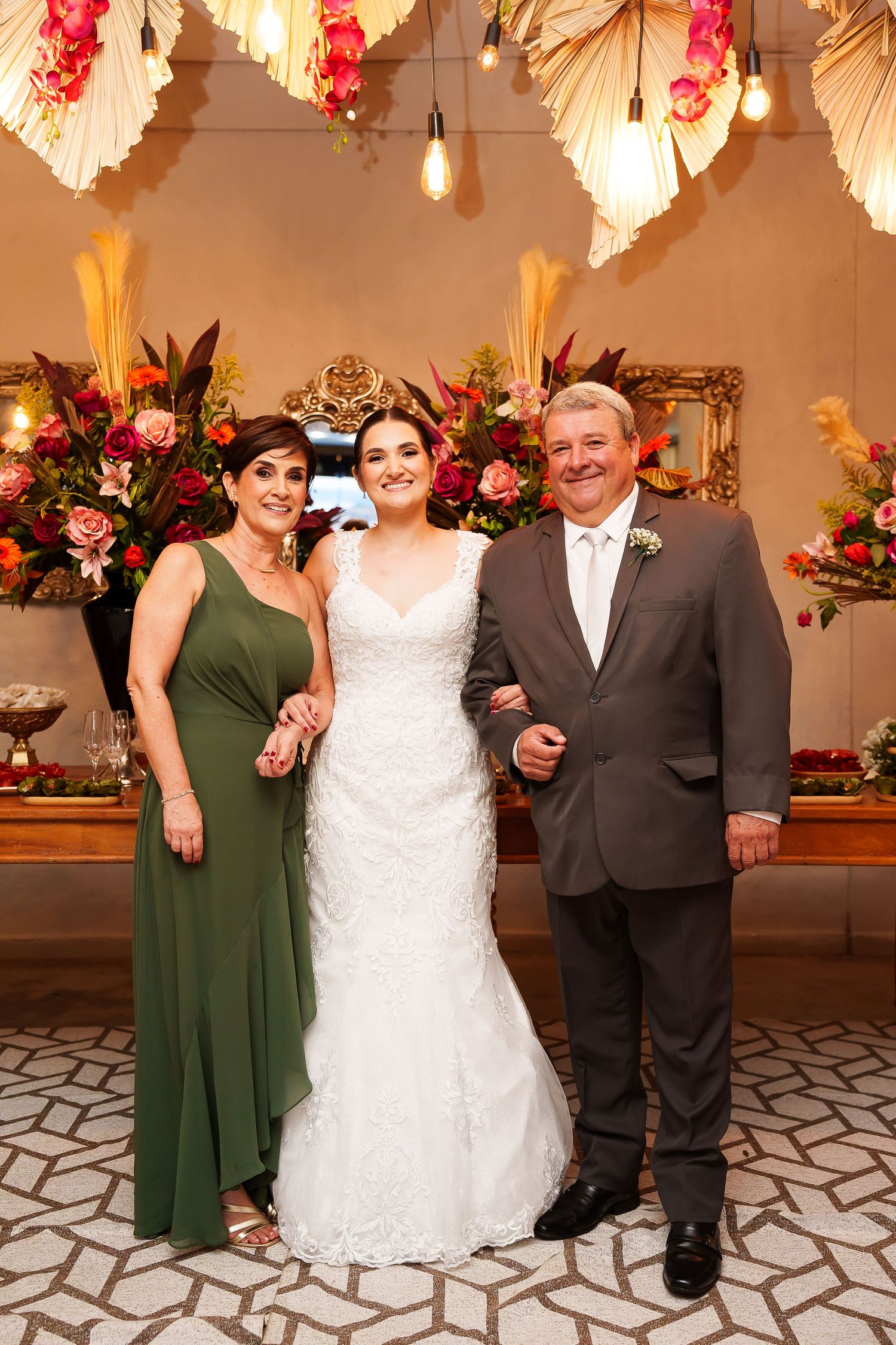 Bride in white lace gown poses with parents in elegant wedding venue with floral decor.
