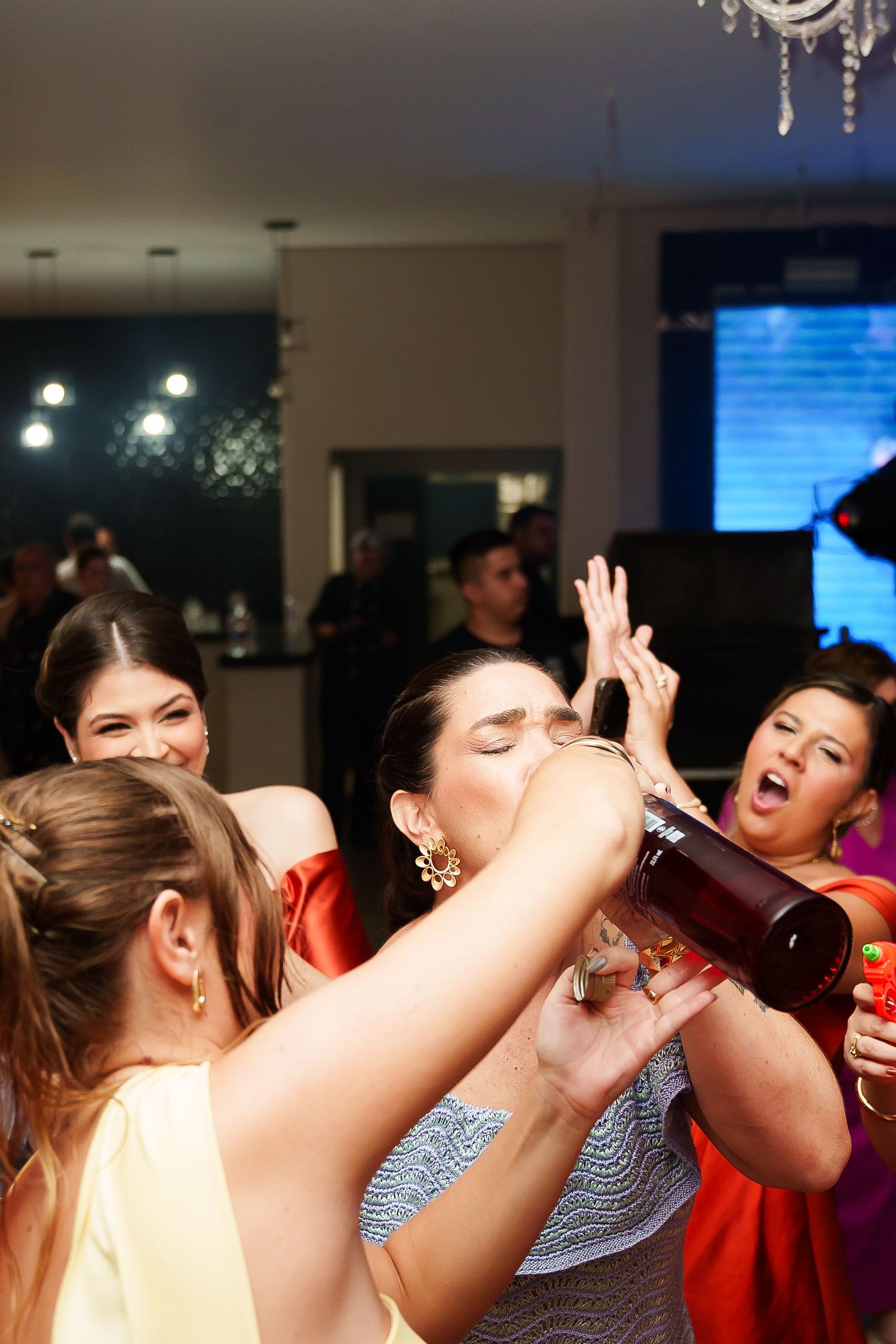 Women celebrating at a party, one drinking from a wine bottle while friends laugh and dance around her.