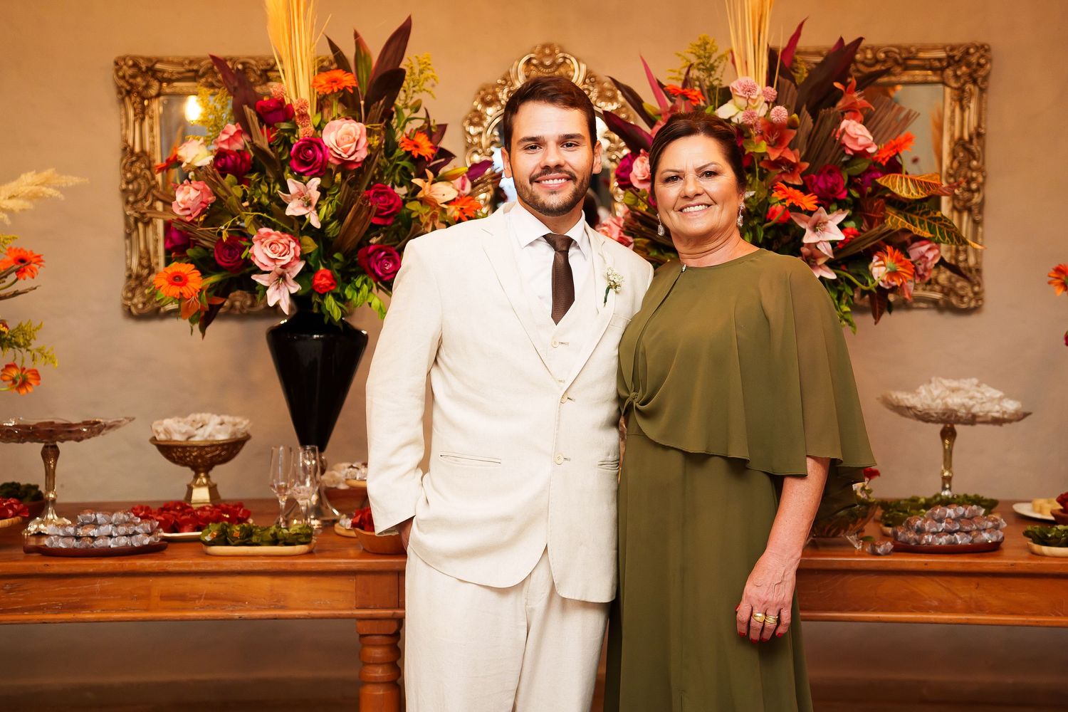 A man in a white suit and woman in olive green dress pose together at an elegant floral wedding reception.