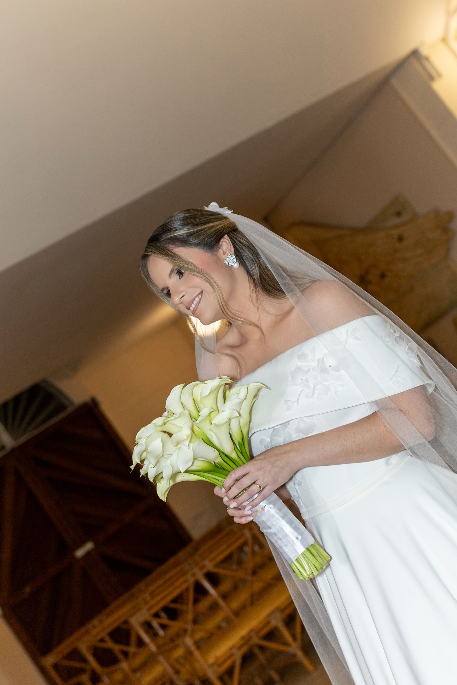 Smiling bride in off-shoulder white gown holding calla lily bouquet with veil, standing indoors.
