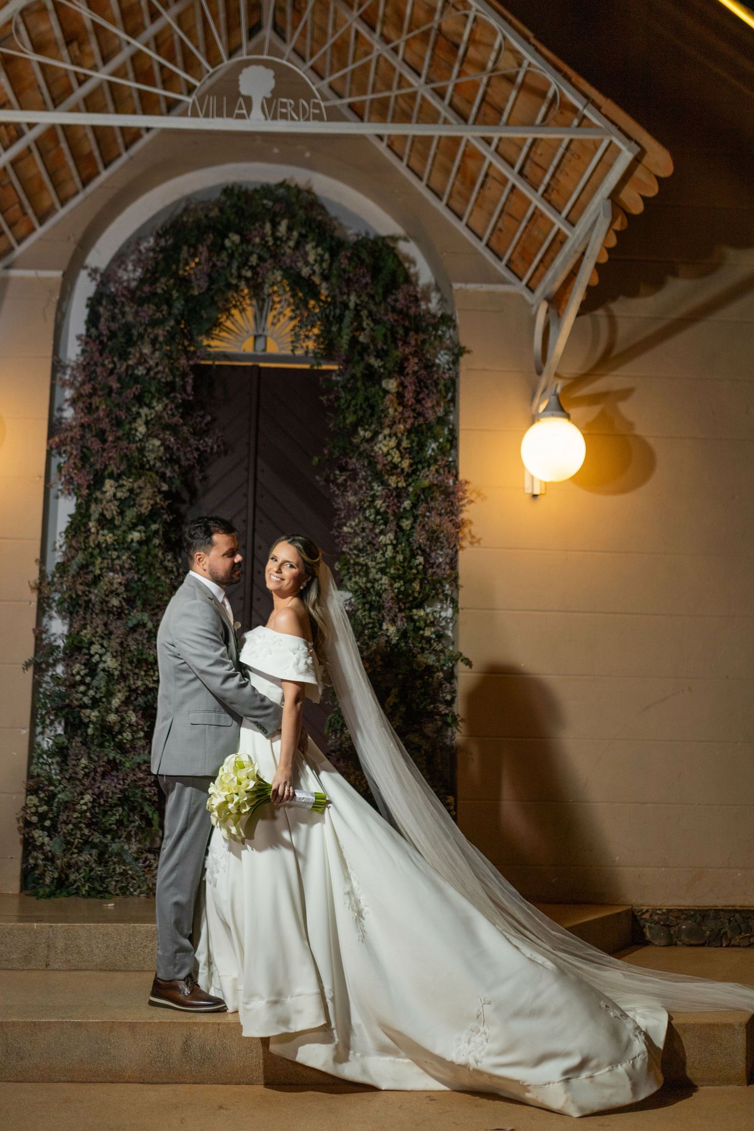 Bride and groom pose on steps before floral-adorned chapel doors at night, bride in elegant gown with long veil.