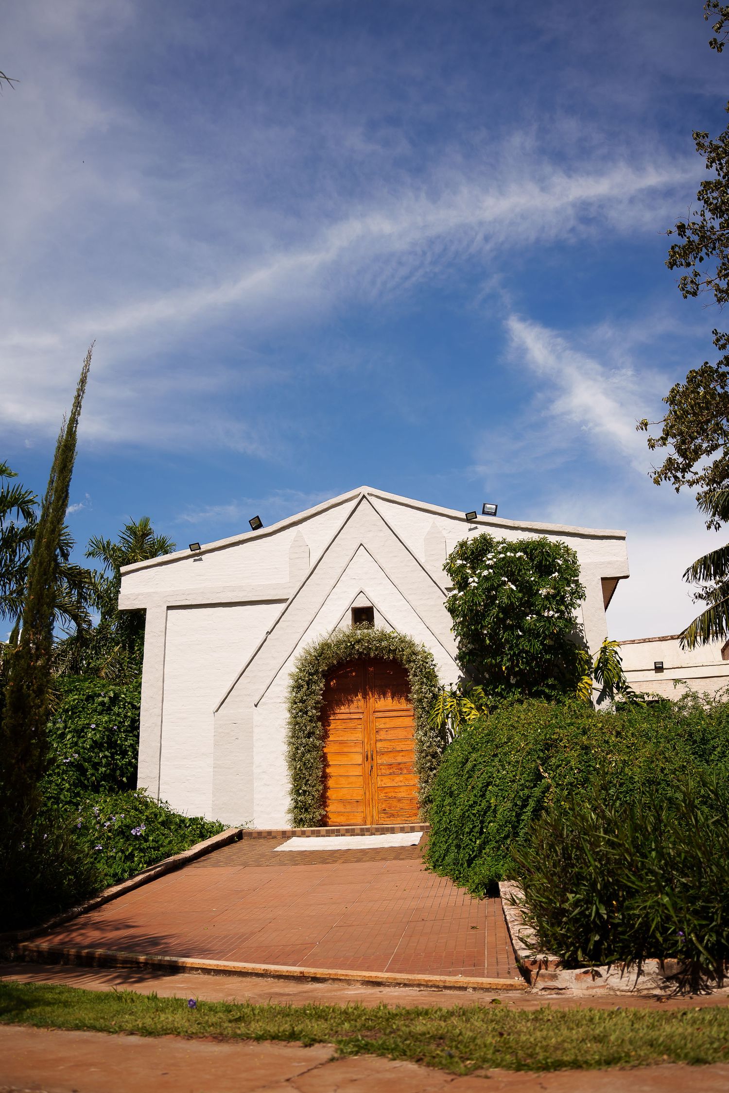 White stucco building with arched wooden door, lush greenery, and brick pathway under blue sky.