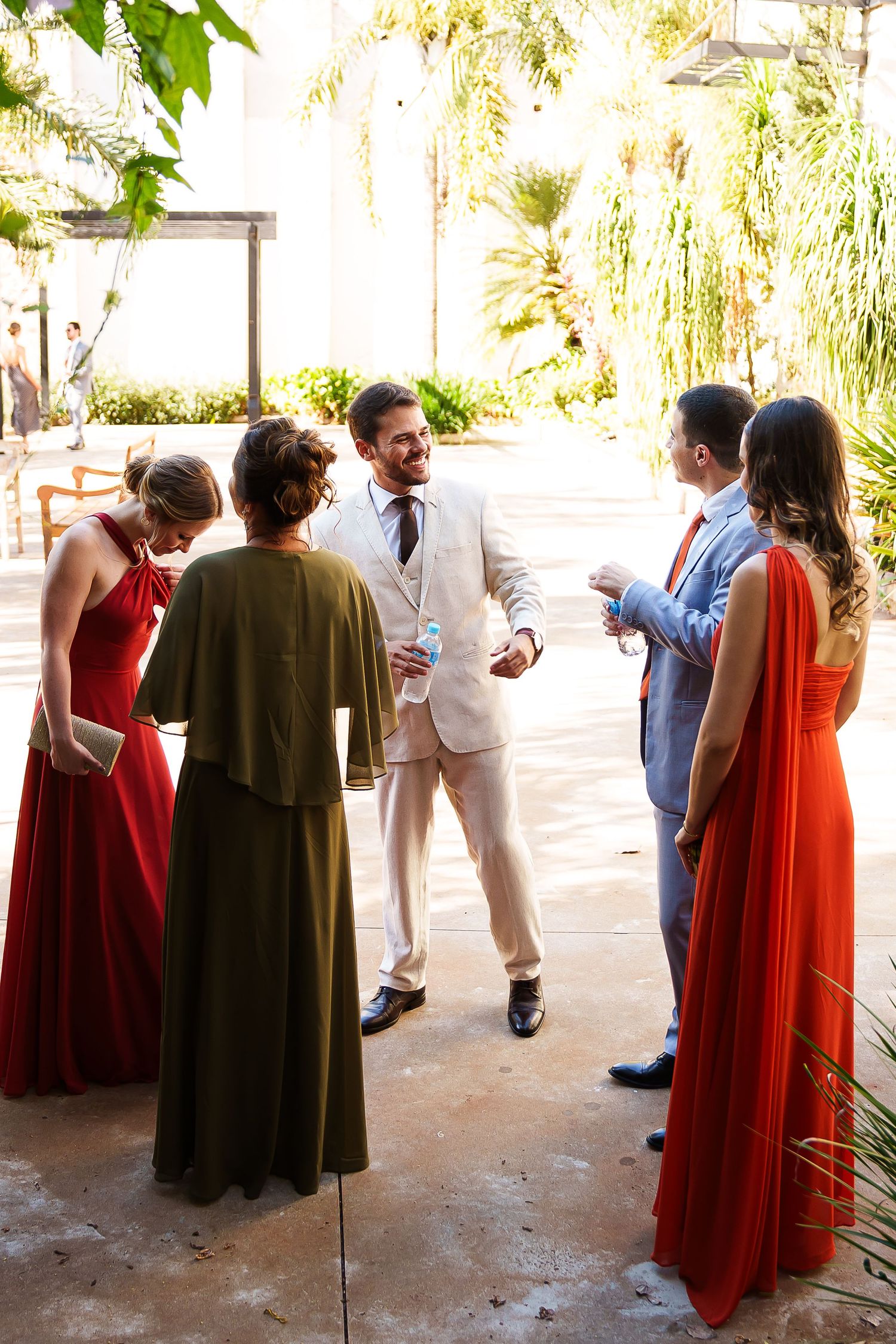 Groom in cream suit mingles with wedding guests in red gowns and formal attire at an outdoor garden venue.
