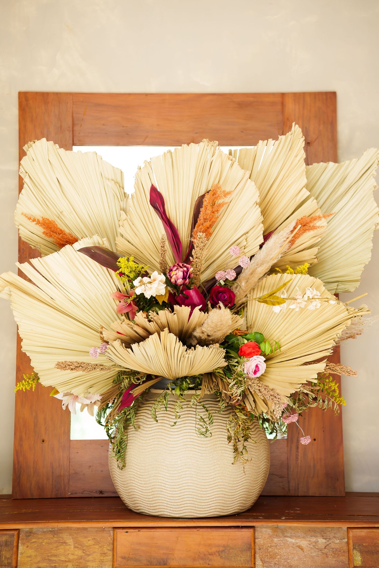 Dried palm leaf and colorful floral arrangement in white vase on wooden dresser with mirror backdrop.