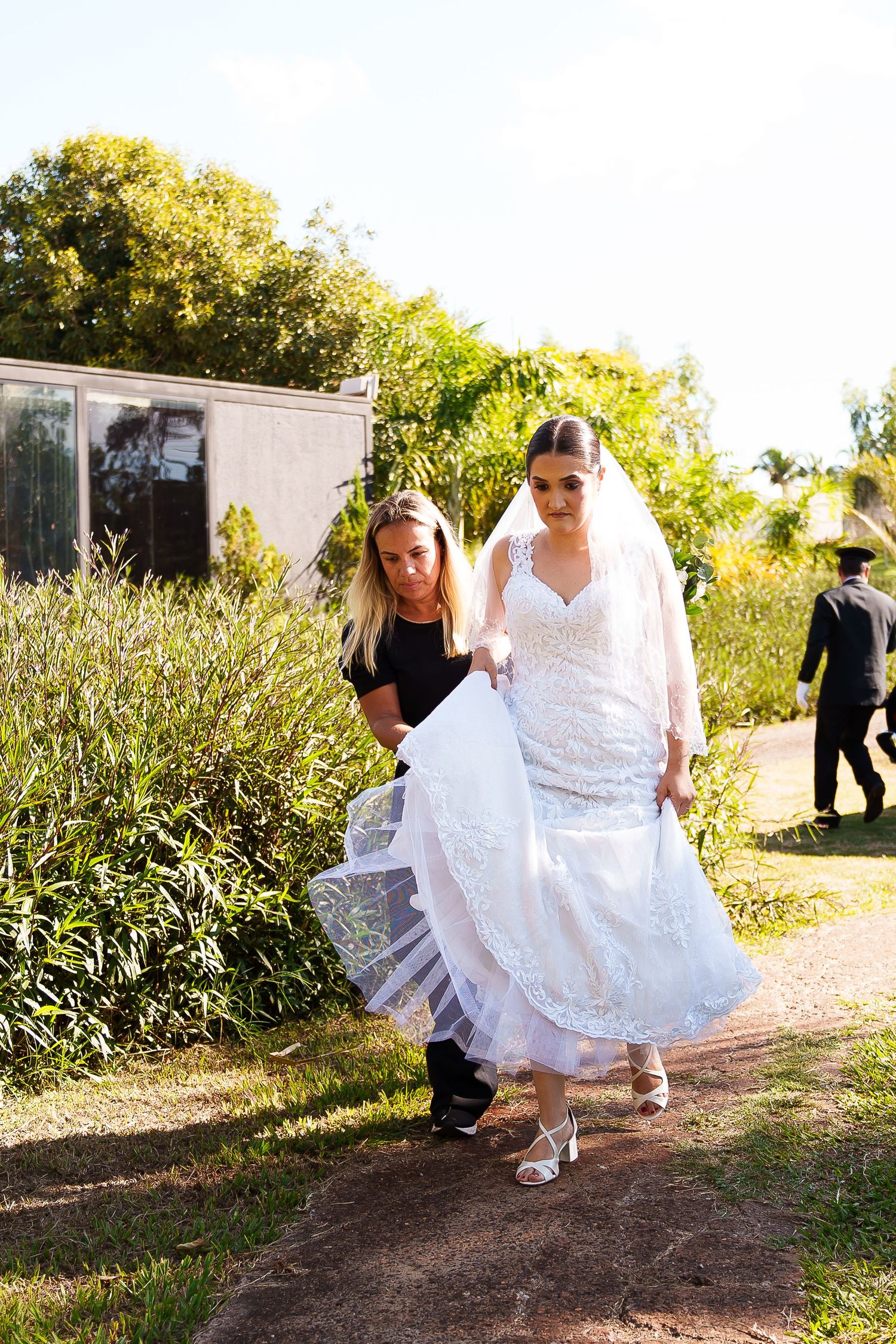 Bride in white lace gown and veil walks outdoors as bridesmaid helps carry her train on a sunny day.