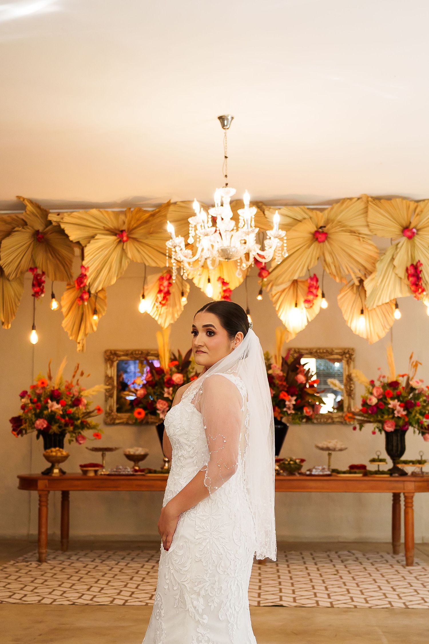 Bride in white lace gown and veil poses before elegant floral backdrop with chandelier at wedding reception.