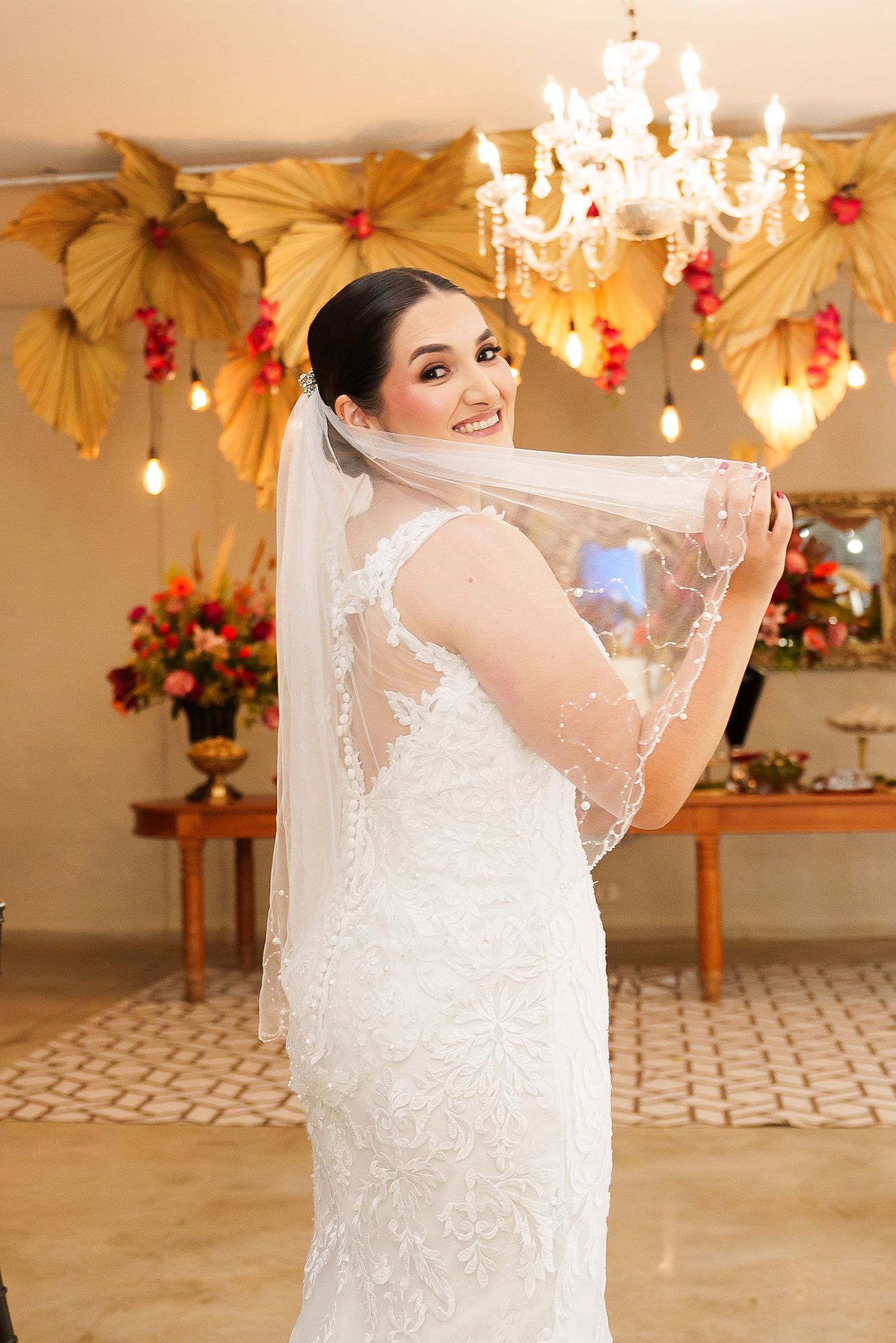 Smiling bride in lace wedding dress holding veil, posing in decorated venue with chandelier and floral arrangements.