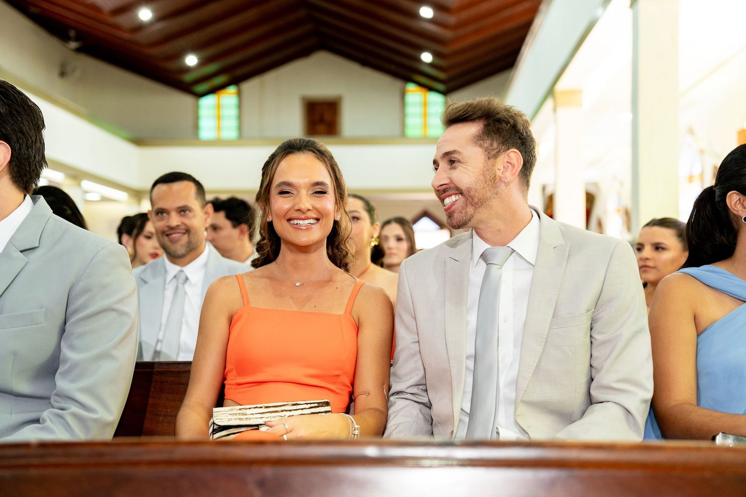 Smiling couple in church pew at wedding ceremony, woman in orange dress, man in beige suit.
