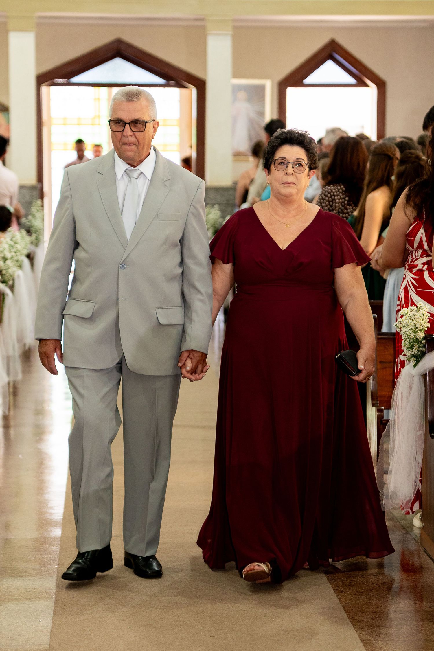 Elderly couple walks down church aisle, man in gray suit, woman in burgundy gown at formal wedding.