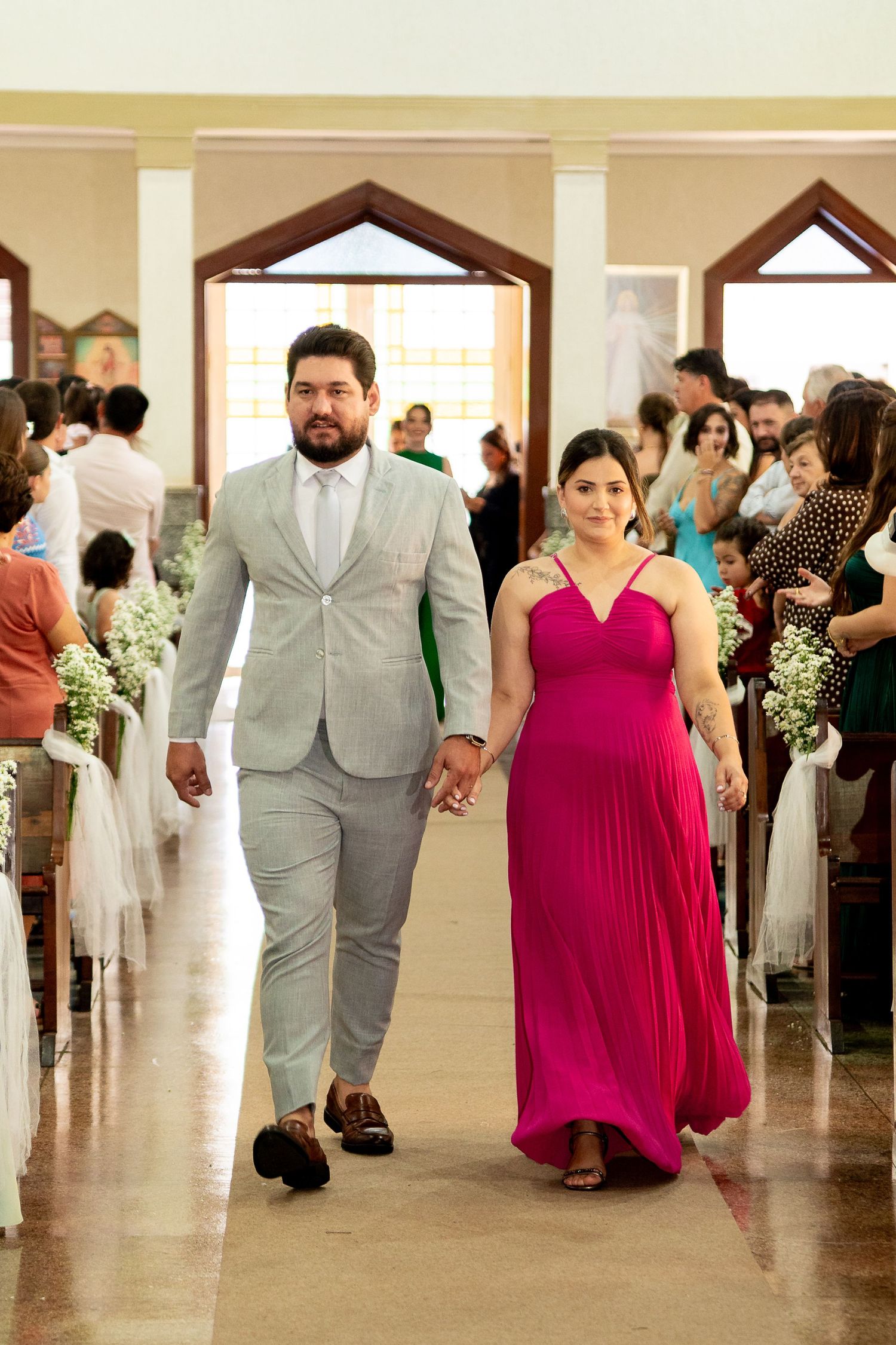 Couple walking down church aisle at wedding, man in gray suit, woman in pink dress, guests seated on pews.