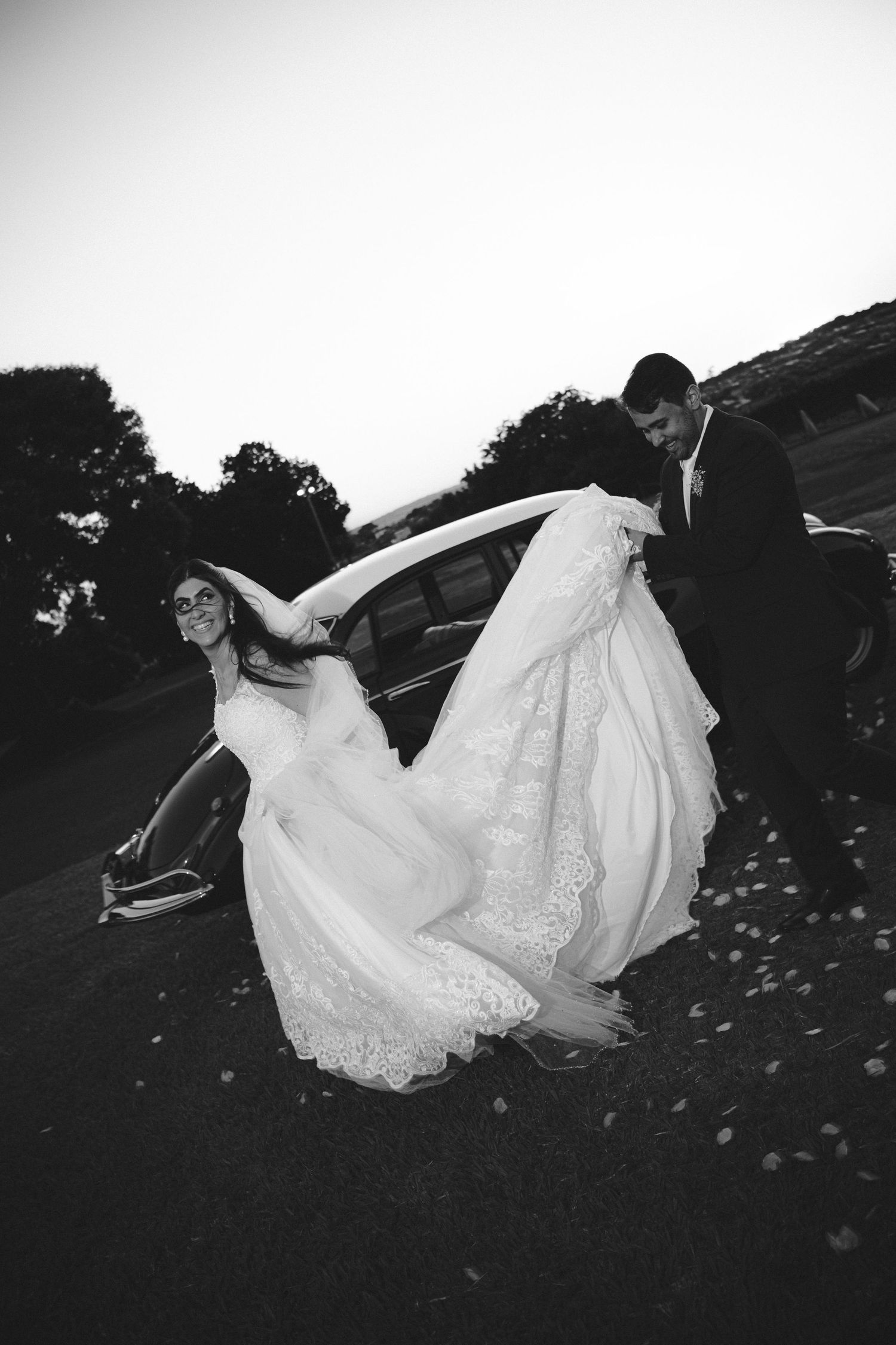 Black and white photo of a bride in a lace gown falling while groom holds her train near a vintage car.