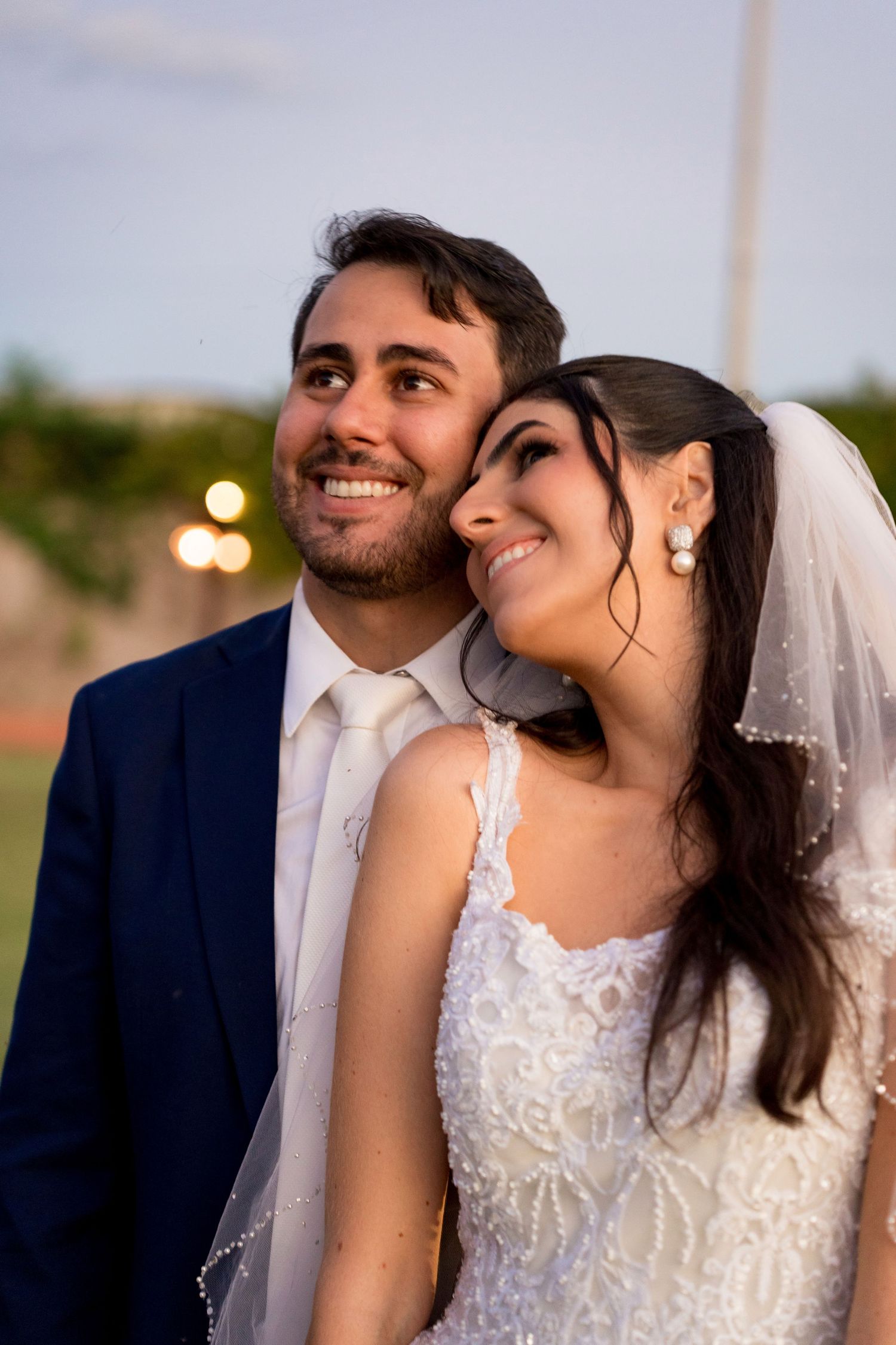 Smiling bride and groom in wedding attire pose closely together outdoors at dusk, with bokeh lights in the background.