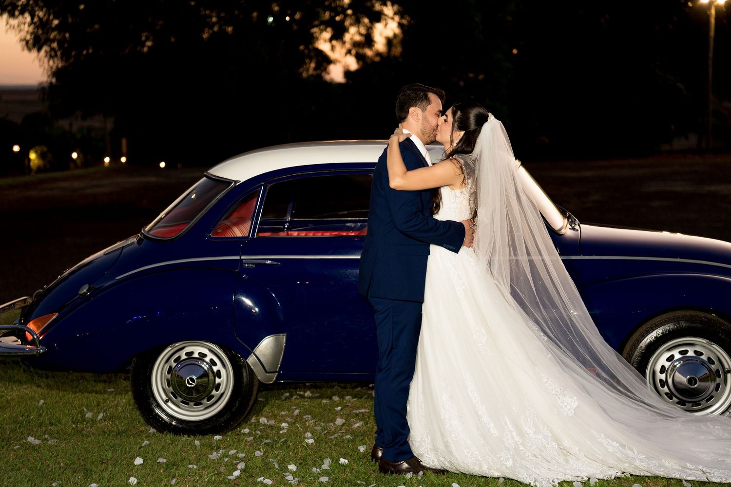 Bride and groom sharing a romantic kiss beside a classic blue vintage car at night during their wedding celebration.