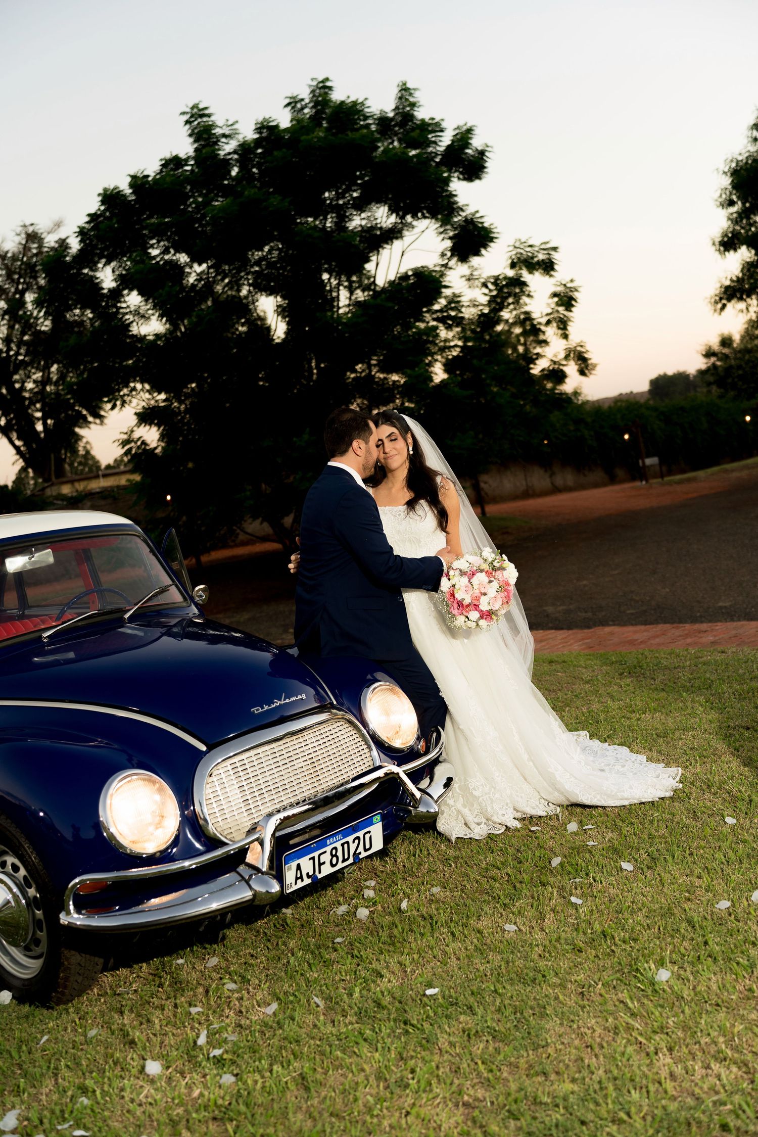 Bride and groom kissing beside a vintage blue car at sunset, bride holding a pink bouquet in a white gown.