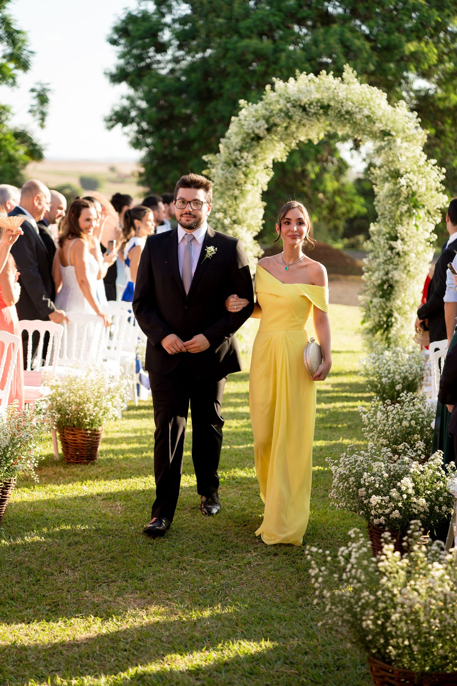 A couple walks down an outdoor wedding aisle, man in dark suit, woman in yellow gown, under a white floral arch.