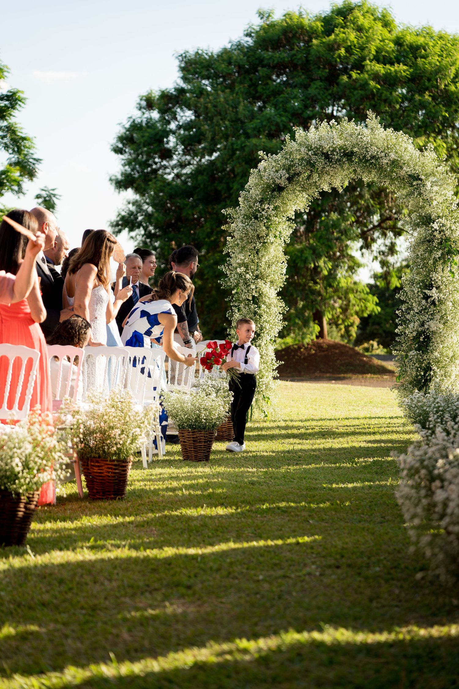 Young ring bearer walks down floral arch-adorned outdoor wedding aisle while guests watch and celebrate.