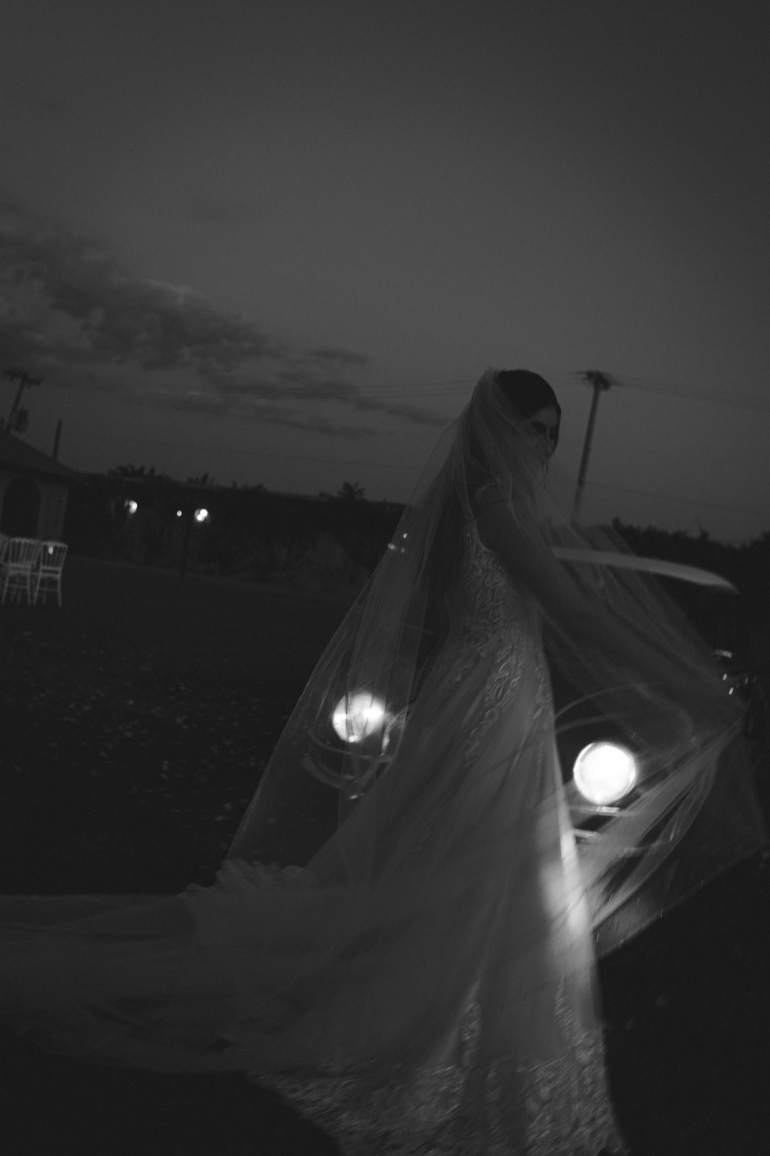 Black and white photo of a bride in a flowing veil standing near car headlights at dusk.