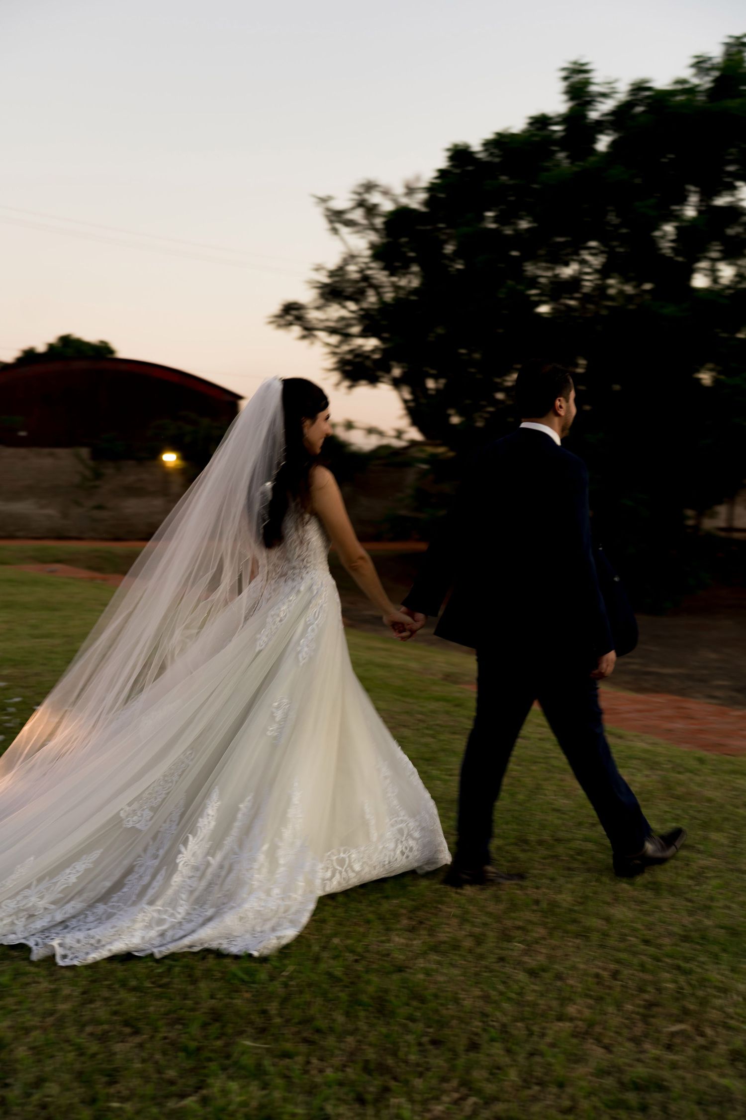 Newlyweds walk hand-in-hand at sunset, bride wearing flowing lace gown with cathedral veil.