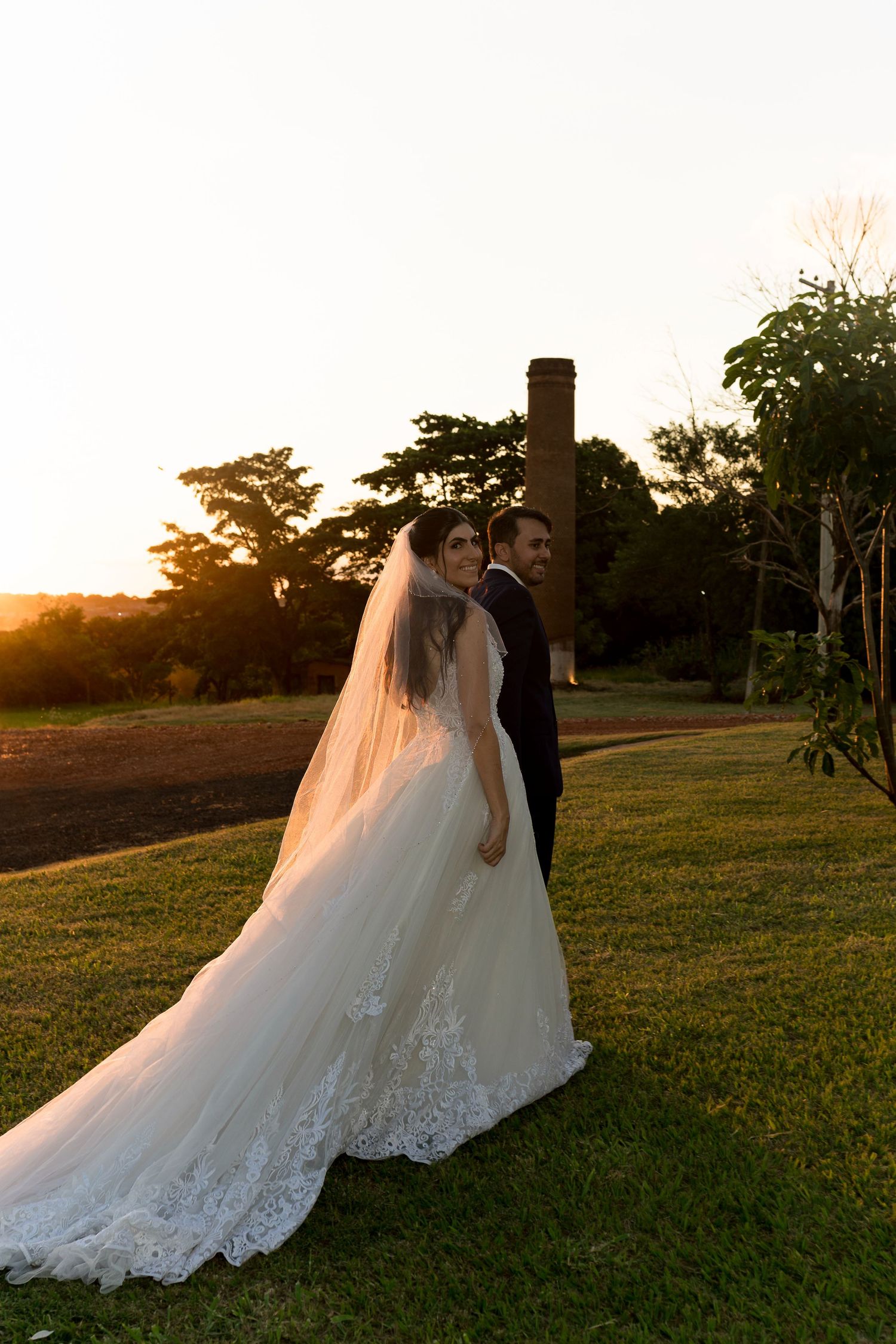 Bride in lace gown with long veil and groom pose on green lawn at golden sunset with trees and brick chimney.