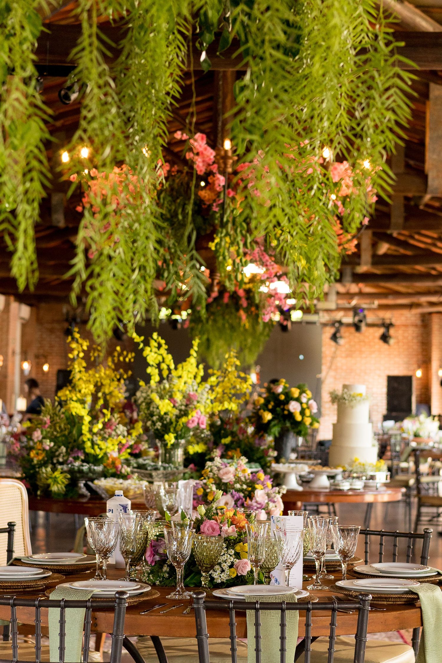 Elegant wedding reception with lush hanging greenery, colorful floral arrangements, and beautifully set round tables in a rustic venue.