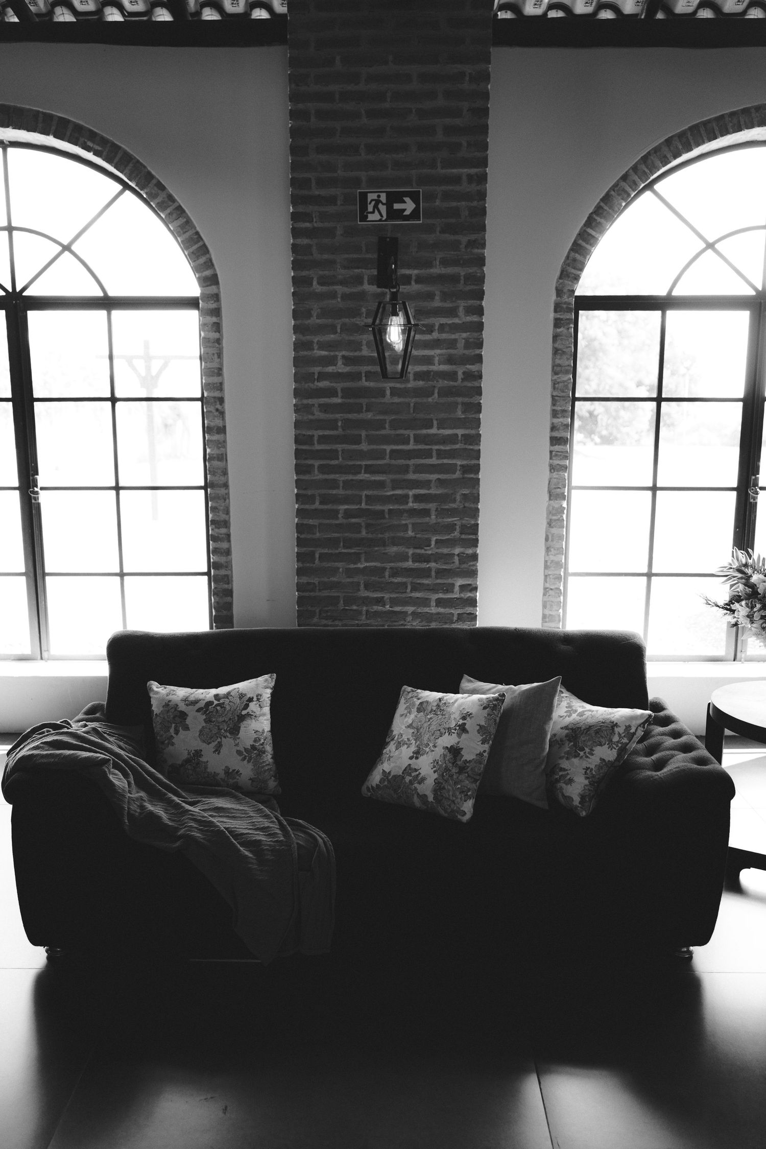 Black and white photo of a tufted sofa with floral pillows between two arched windows and a brick column with wall sconce.