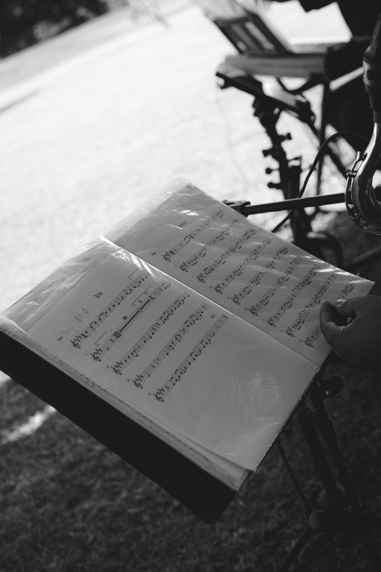 Black and white photo of sheet music on a music stand outdoors, with instruments visible in the background.