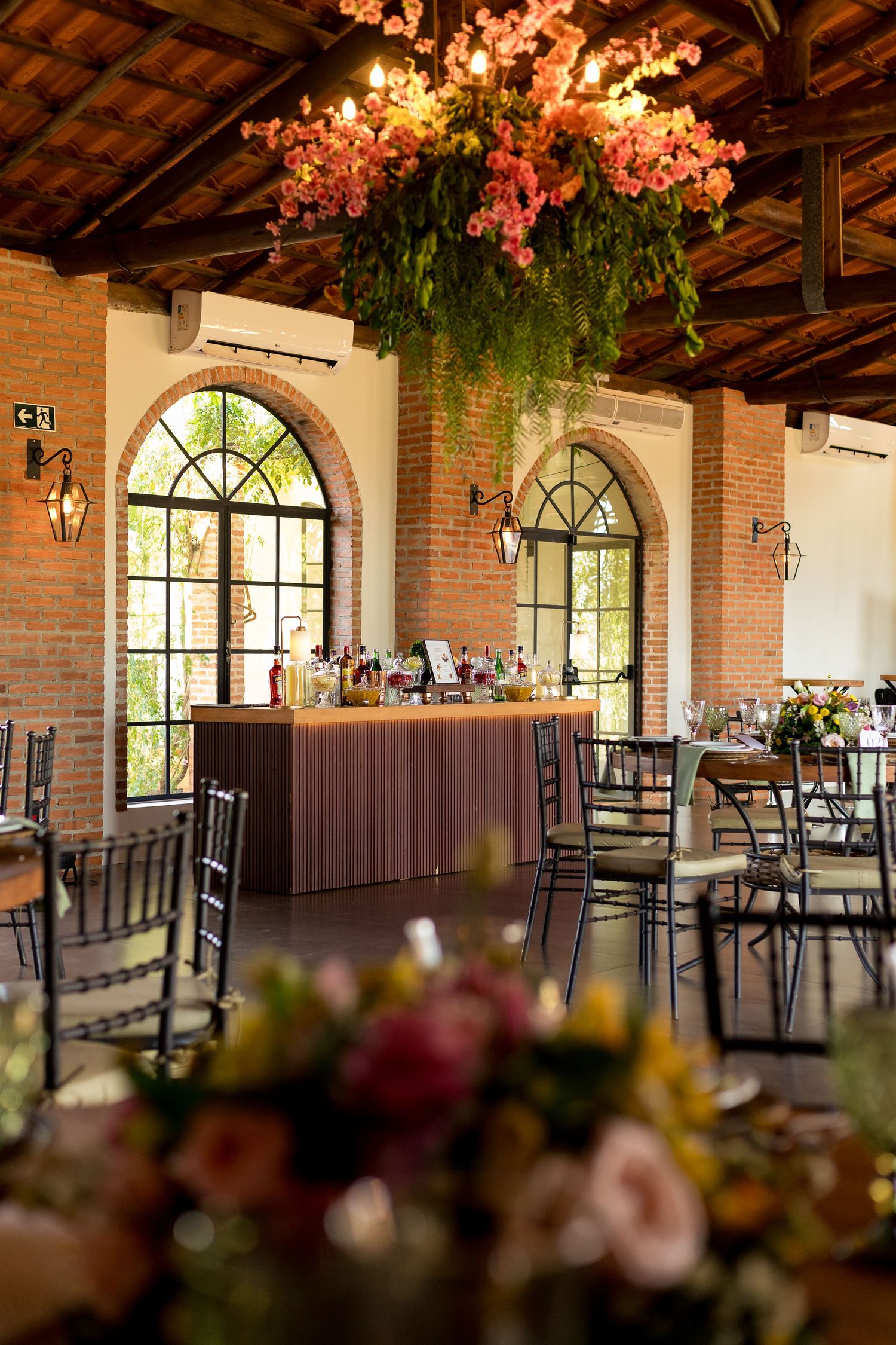 Elegant event venue with brick walls, arched windows, floral ceiling installation, and decorated tables for a wedding reception.