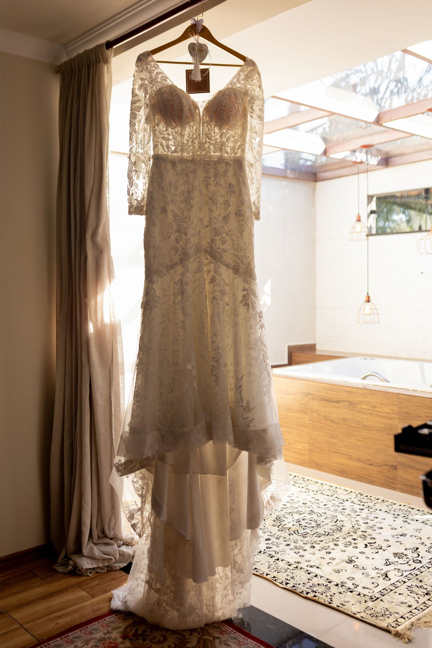 Elegant lace wedding dress hanging on a hanger in a sunlit bridal suite with hardwood floors.