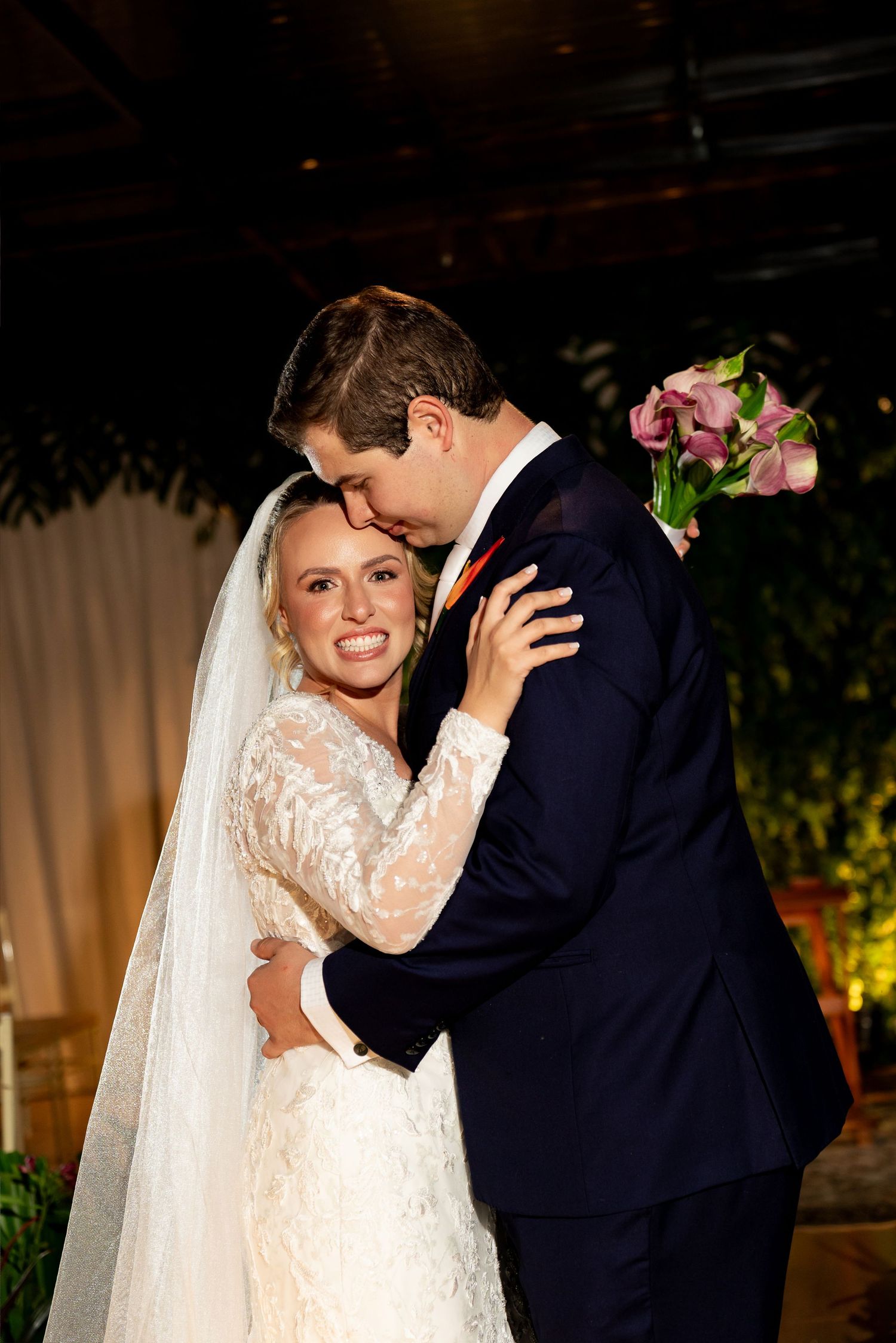 Bride in lace gown and veil embraces groom in navy suit, sharing a tender moment at their wedding reception.