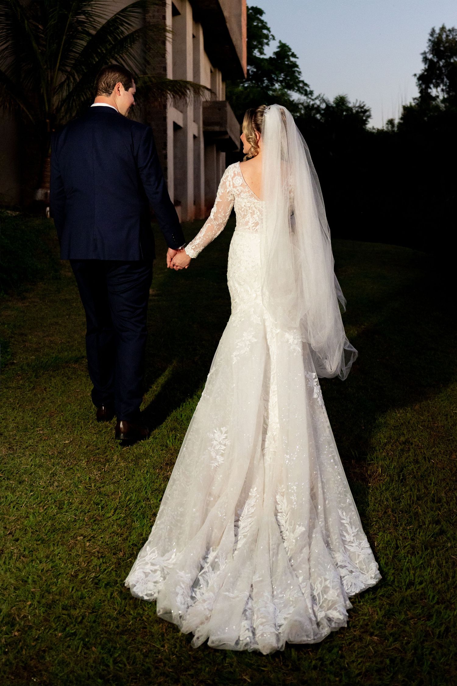 Bride in lace mermaid gown with veil holds groom's hand walking outdoors at golden hour wedding.